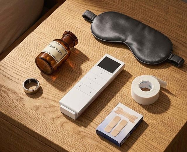 Sleepmaxxing essentials flat lay including magnesium supplements, smart ring, blackout shade remote, mouth tape, and silk sleep mask on oak bedside table