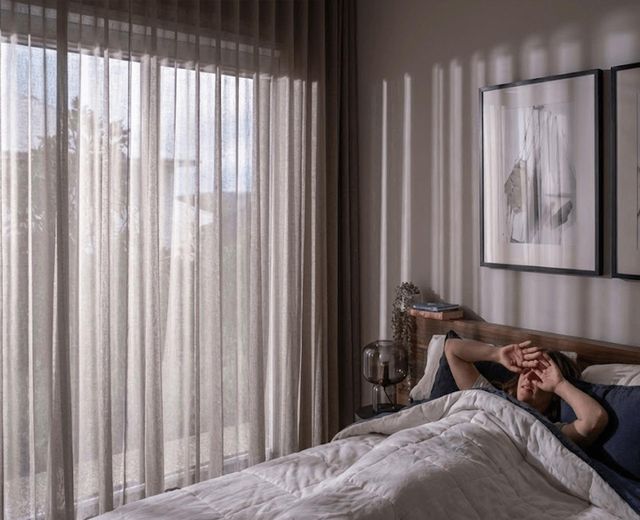 Woman disturbed by morning light leaking through sheer curtains in bedroom