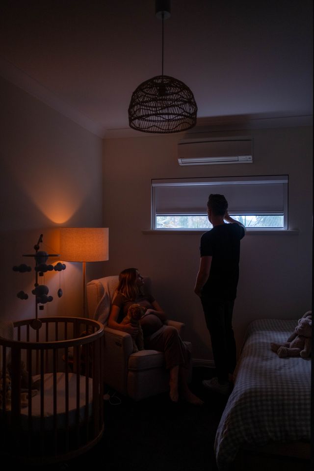 Parents in a dim nursery with baby, father checking window shade for light leaks while mother holds infant