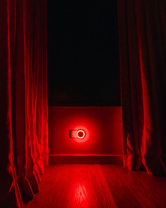 Dark bedroom with dramatic red glow from a circular LED night light between two open floor-length curtains casting red tones across wooden floors