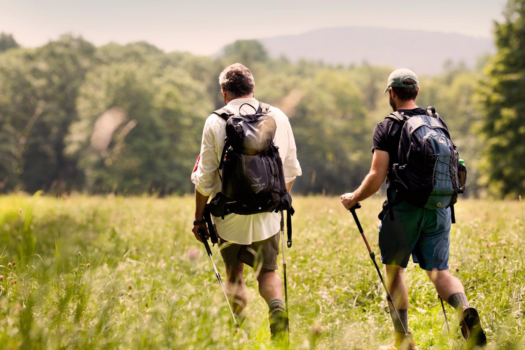 two men hiking through green field with hiking poles and backpacks on