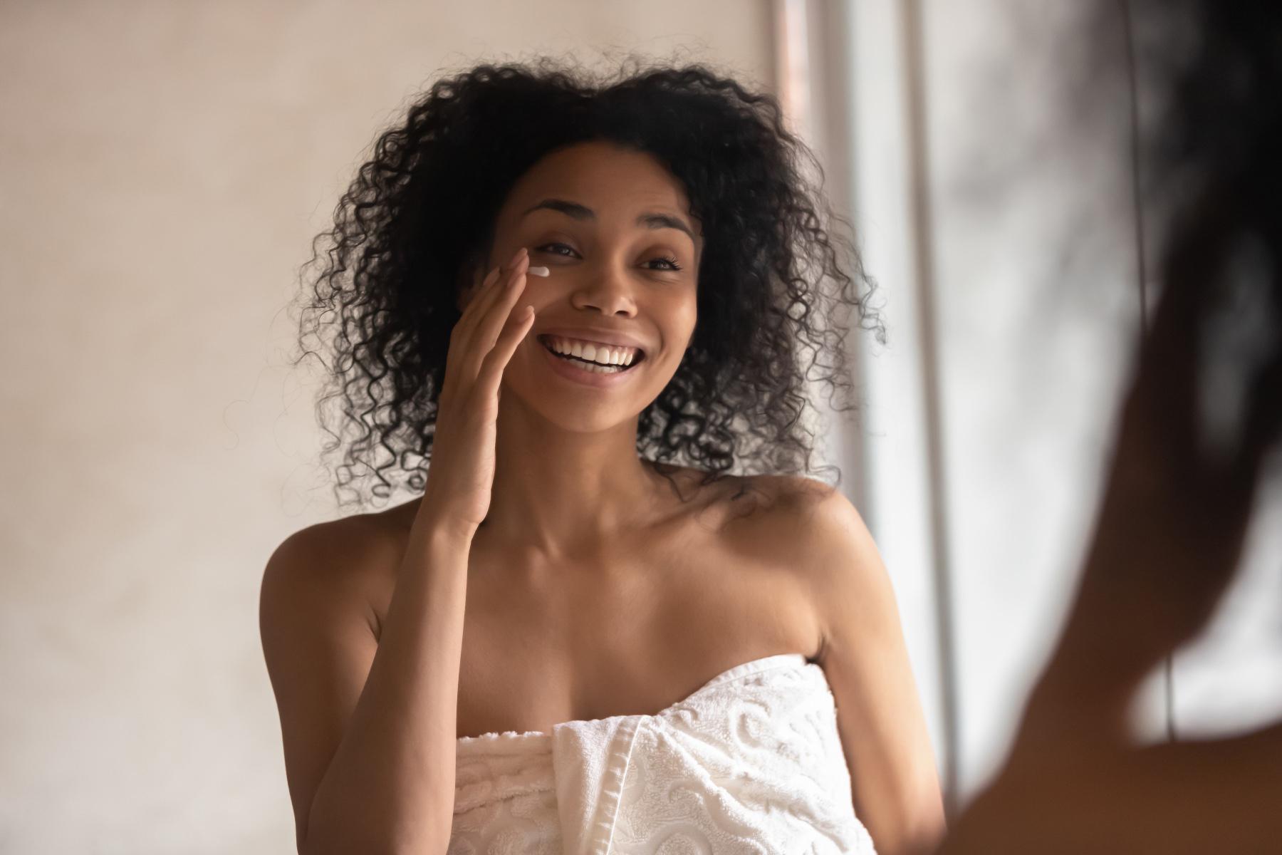 A woman wrapped in a towel applying lotion to her under eyes.