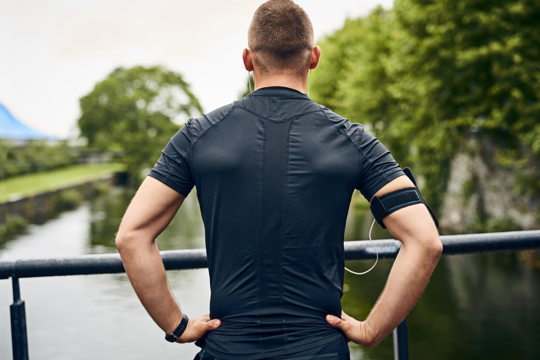 man's back up close as he stands in a black shirt with hands on hips taking a break after working out to look out over bridge