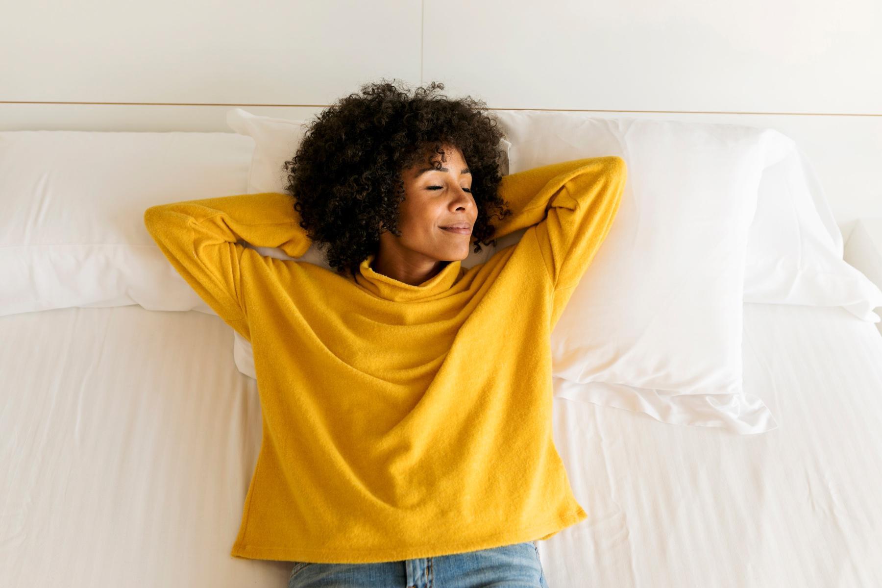 Woman in bright yellow shirt laying on bed on her back with her arms behind her head, smiling with eyes closed.