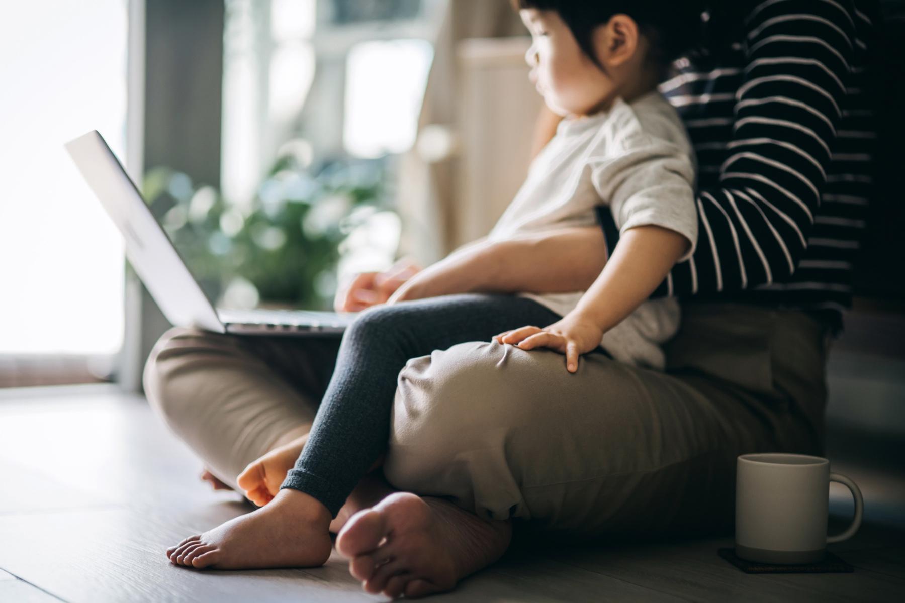 Woman sitting on ground working on laptop with toddler sitting in lap.