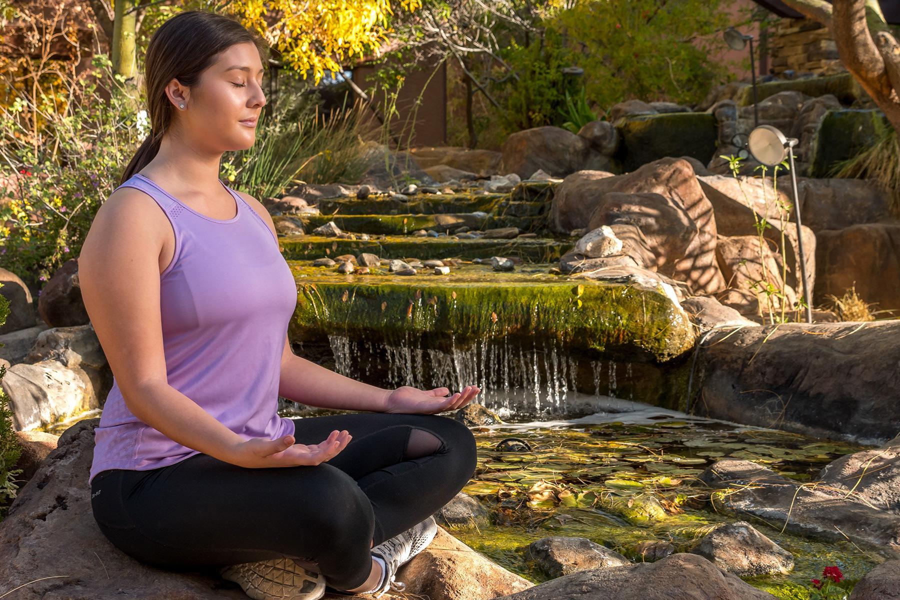 Young woman sitting by outdoor water feature meditating with eyes closed.