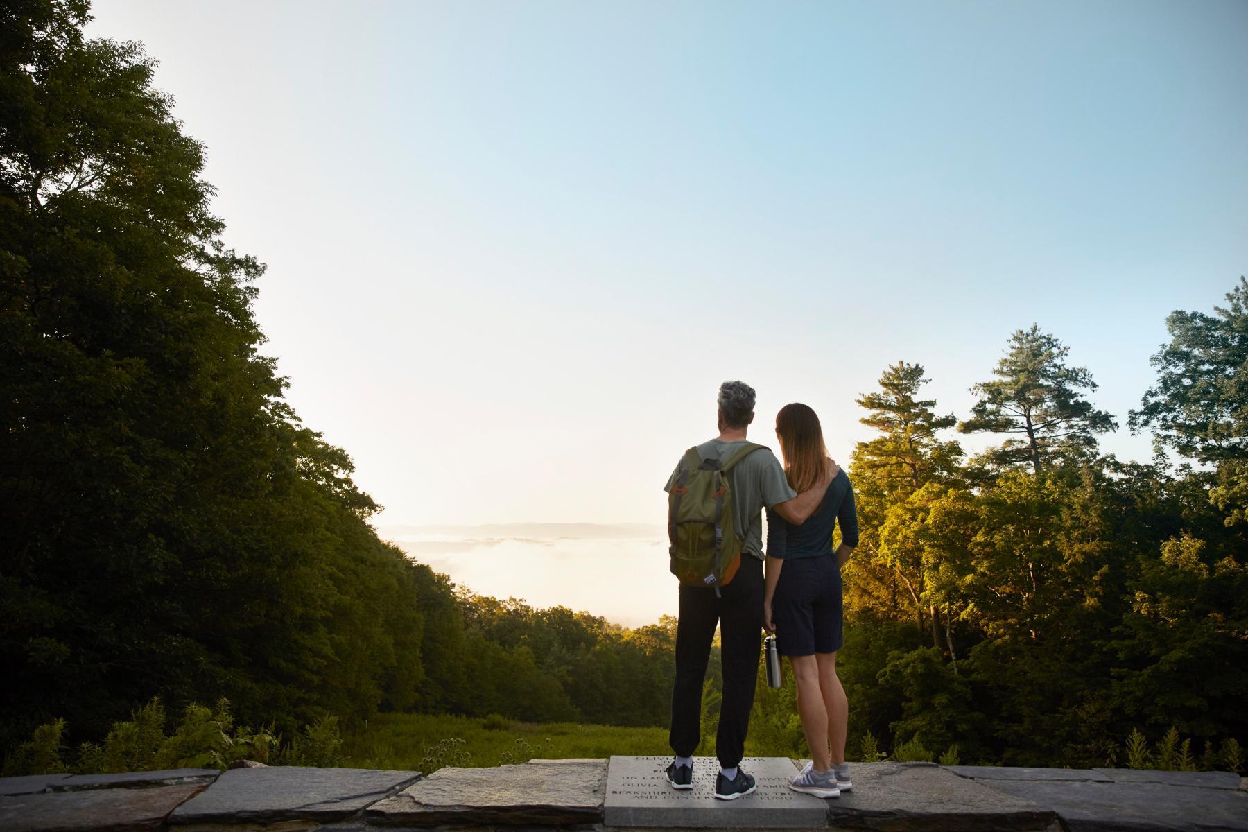 View from behind of man and woman standing at scenic overlook with man's arm around woman.