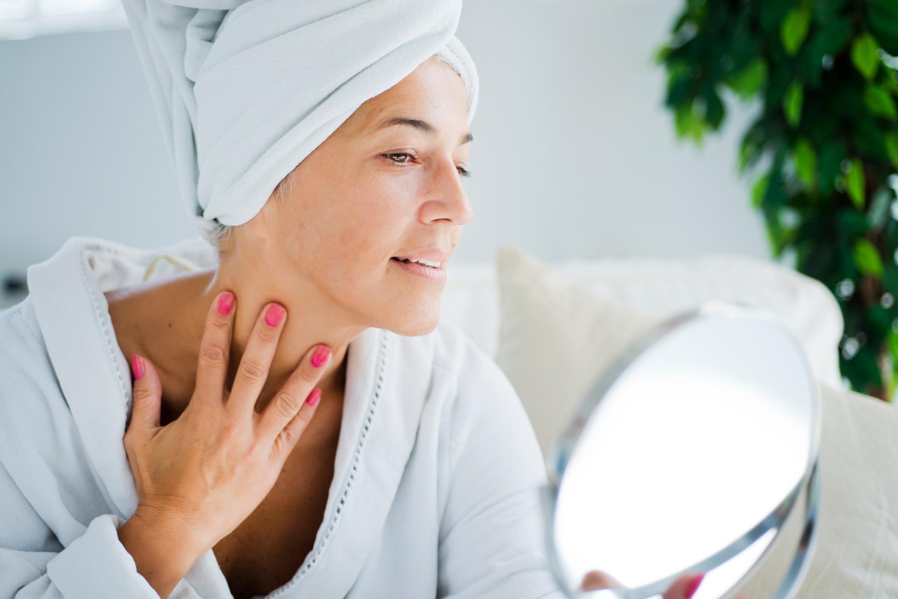 A woman in a robe and her hair in a towel looking at her neck in a handheld mirror.
