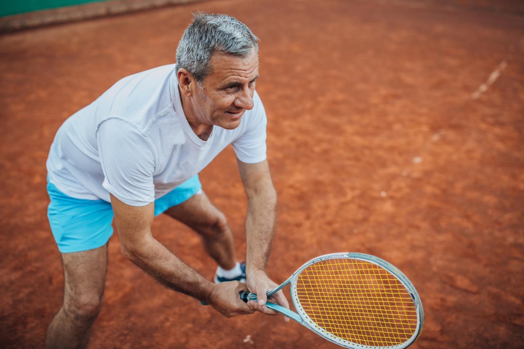 Close-up of an older man holding tennis racket on clay court.
