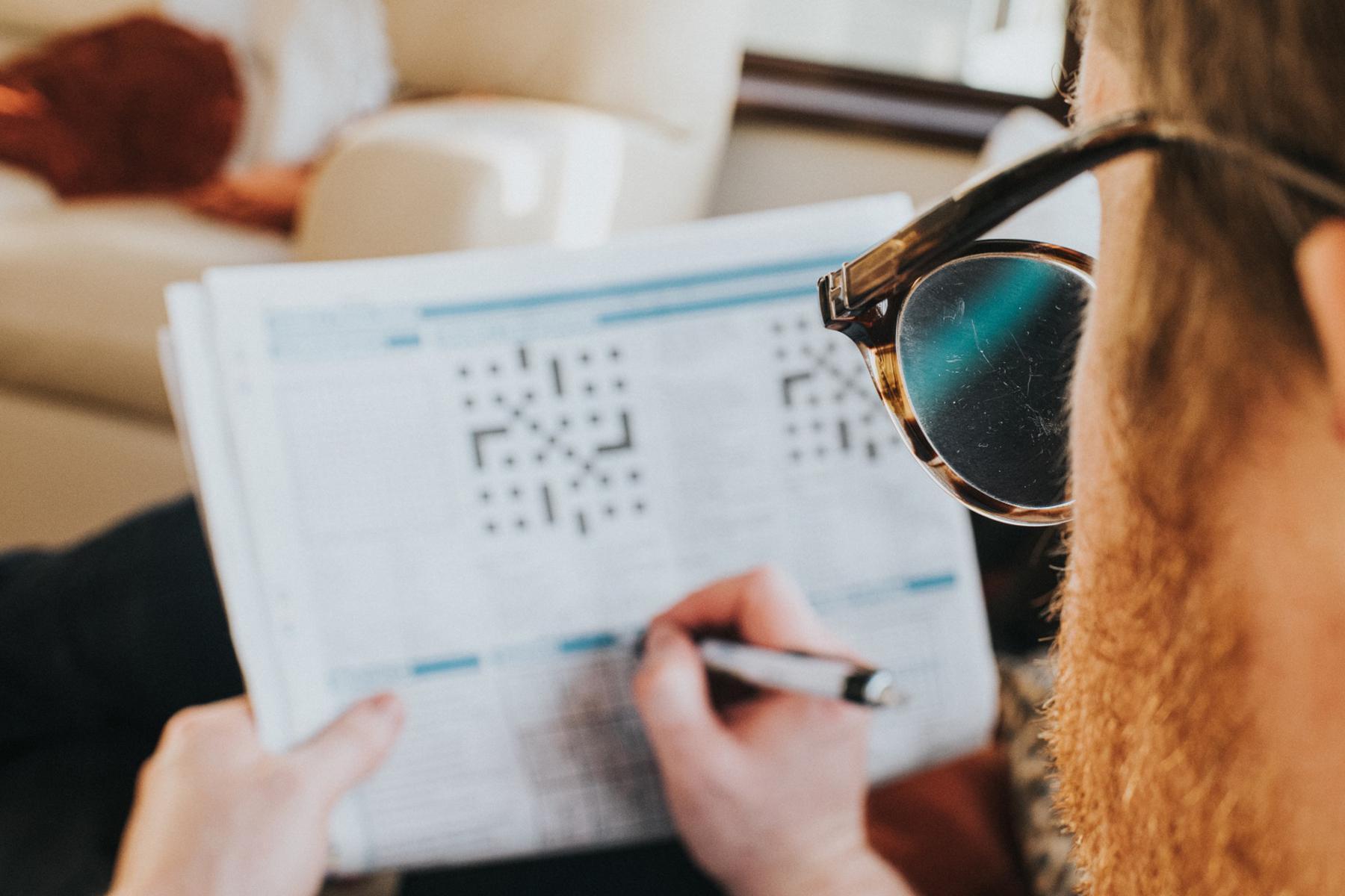 Looking over the shoulder of a man with glasses on doing a crossword puzzle in the newspaper while sitting on a couch.