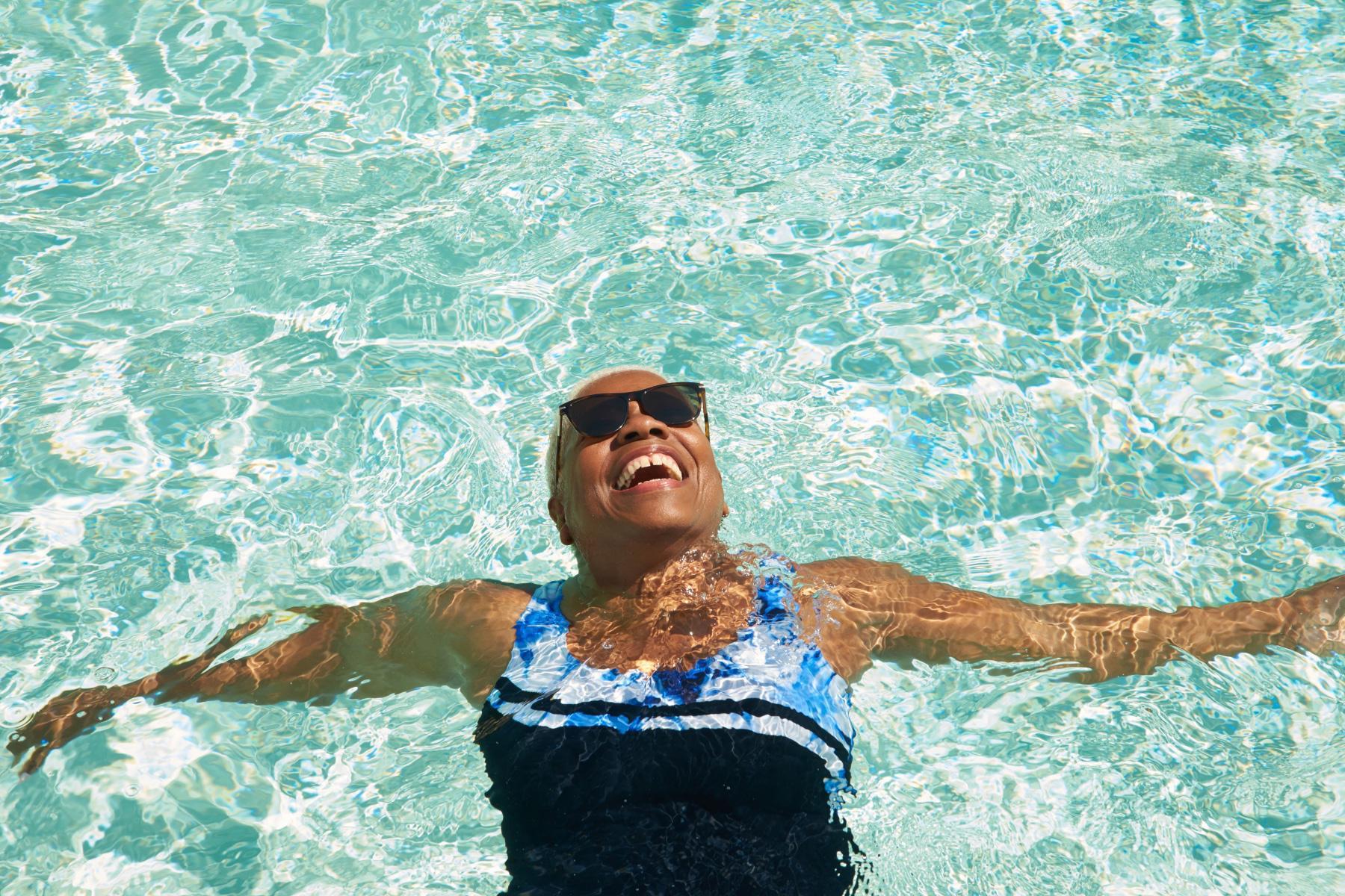 Woman floating on back in pool with sunglasses on smiling at sky.