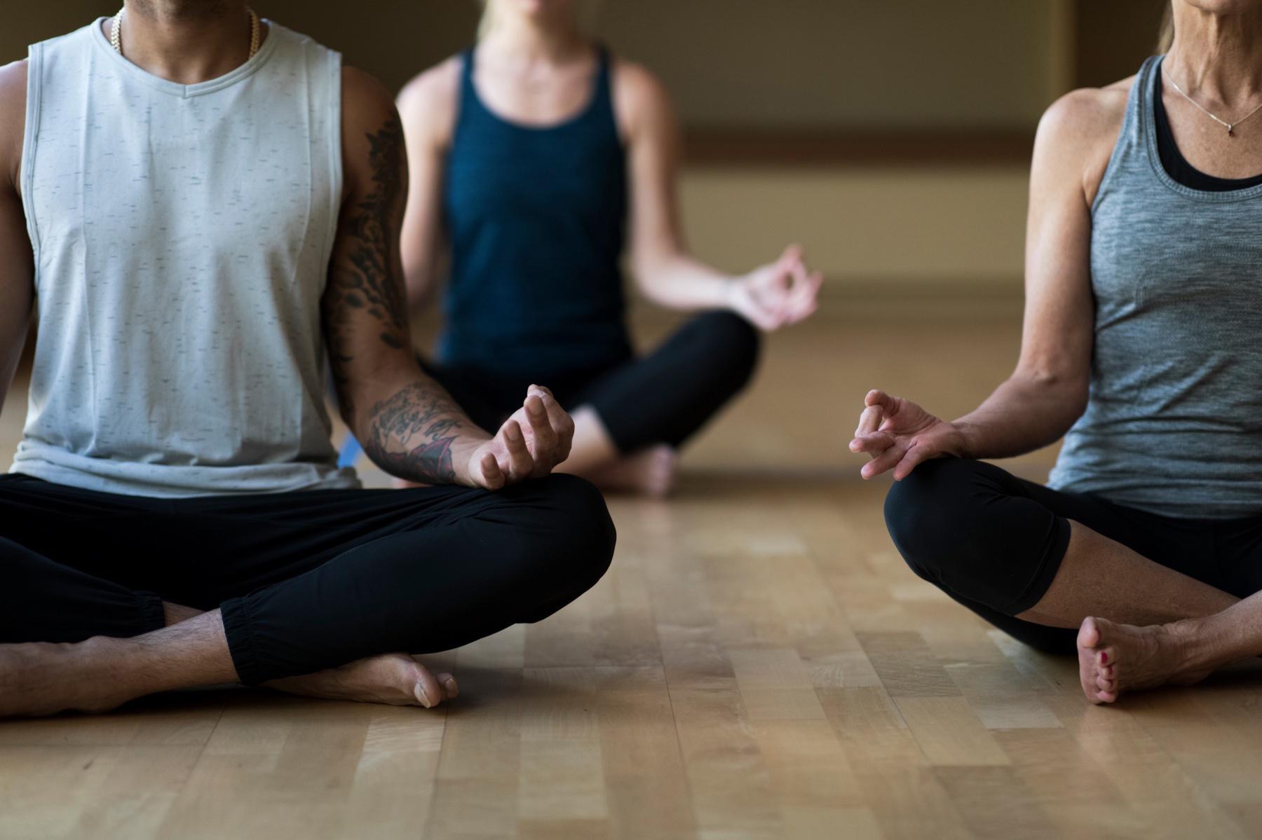 Three people shown from the neck down in a yoga studio sitting in a cross-legged meditation pose.