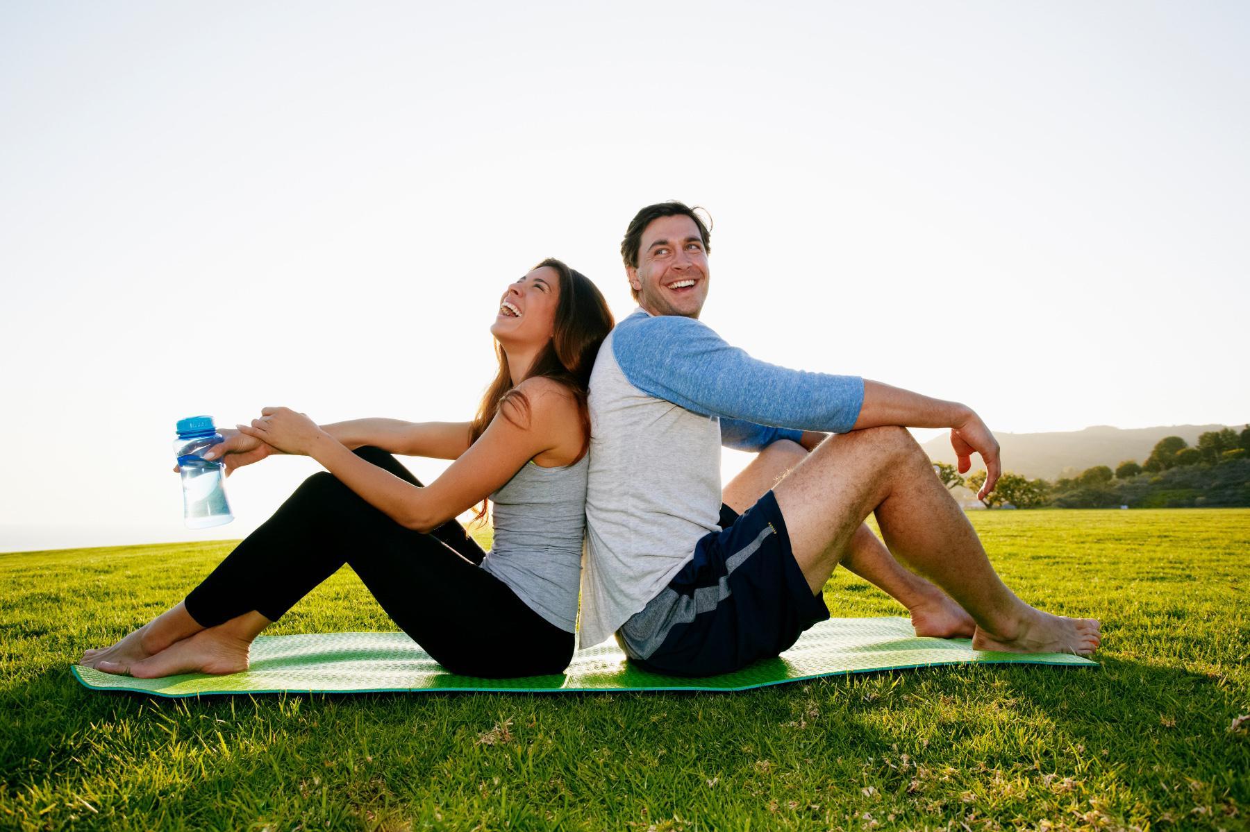man and woman sitting back to back on yoga mat outside laughing and looking up