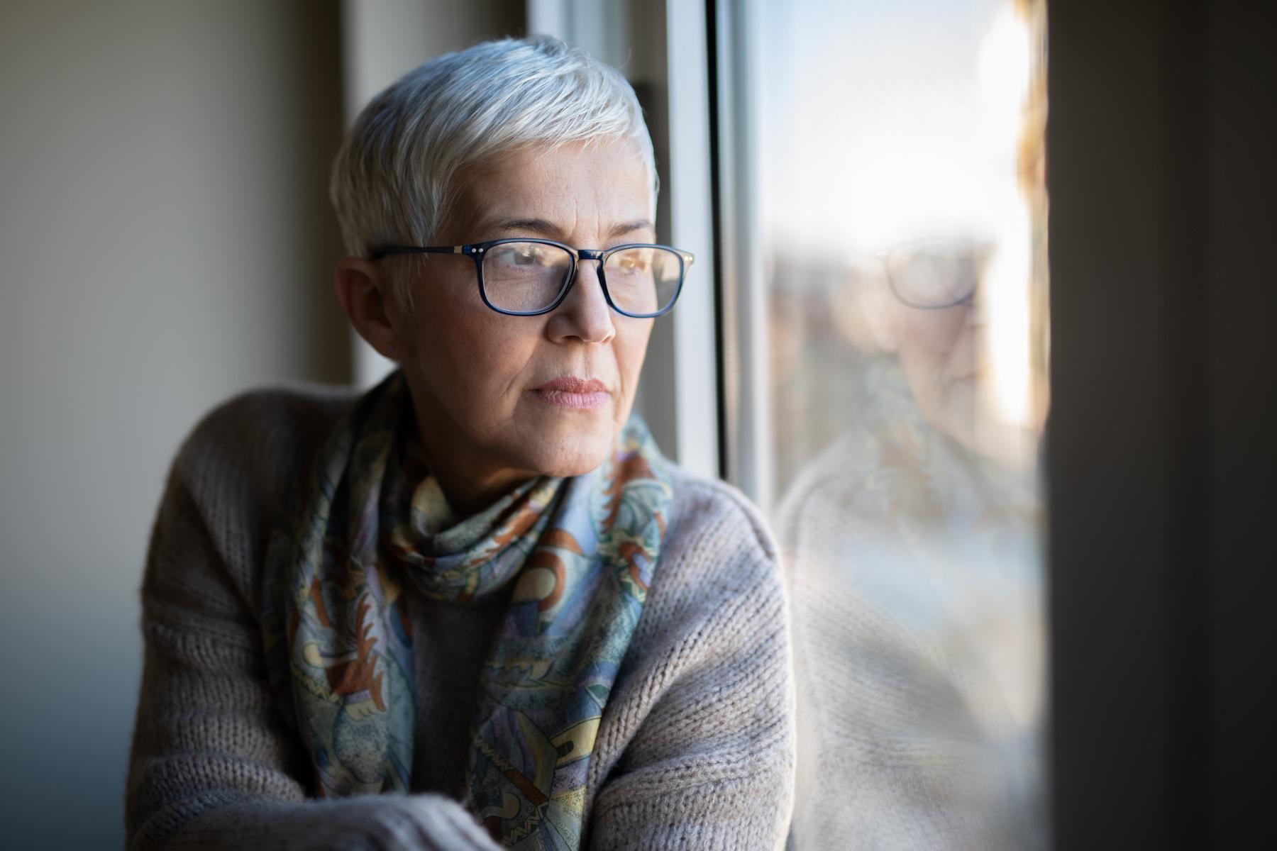 Older woman with glasses sitting up against window looking outside.