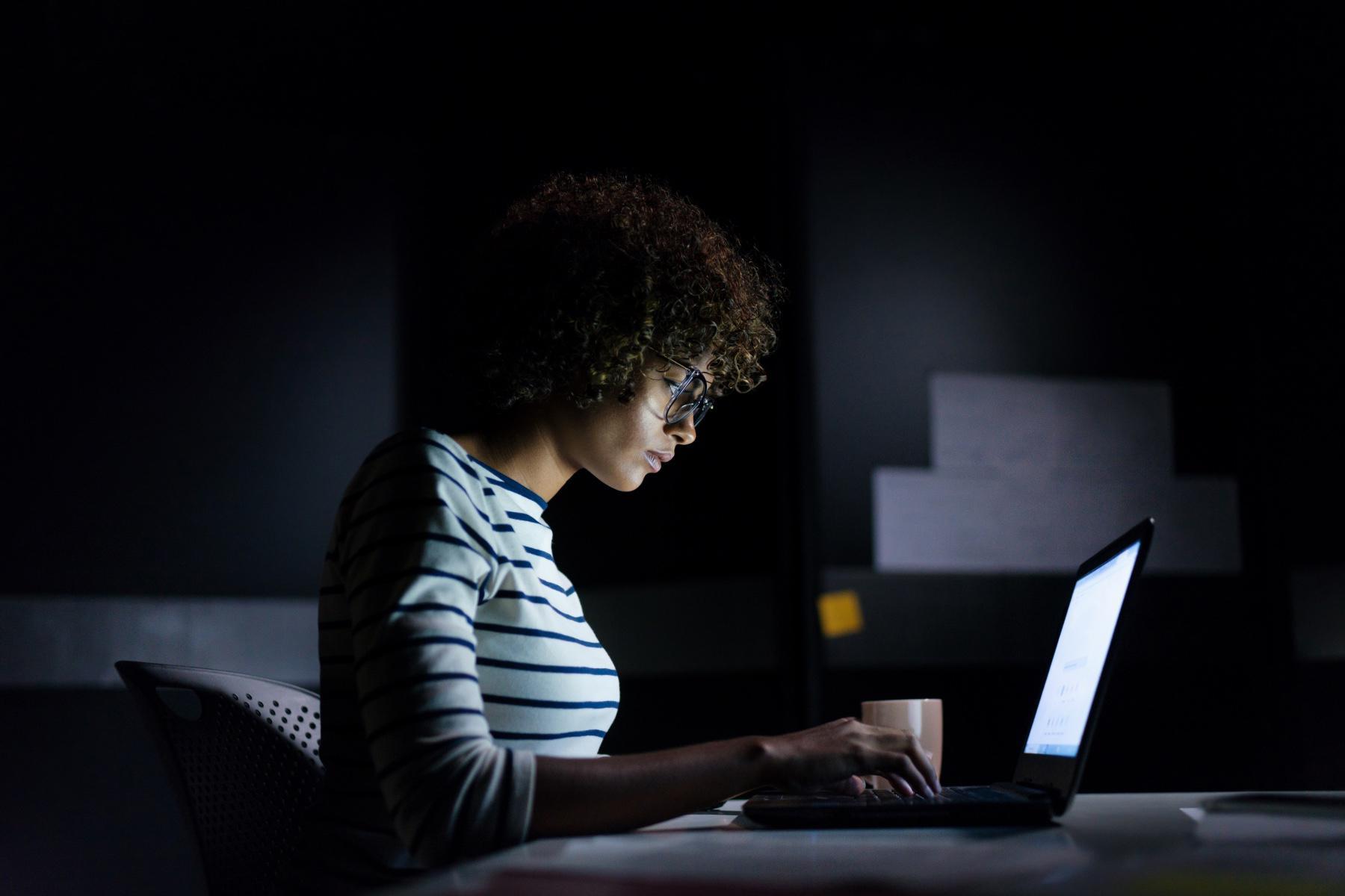 Side view of woman sitting at a table in a dark room looking at her laptop screen.