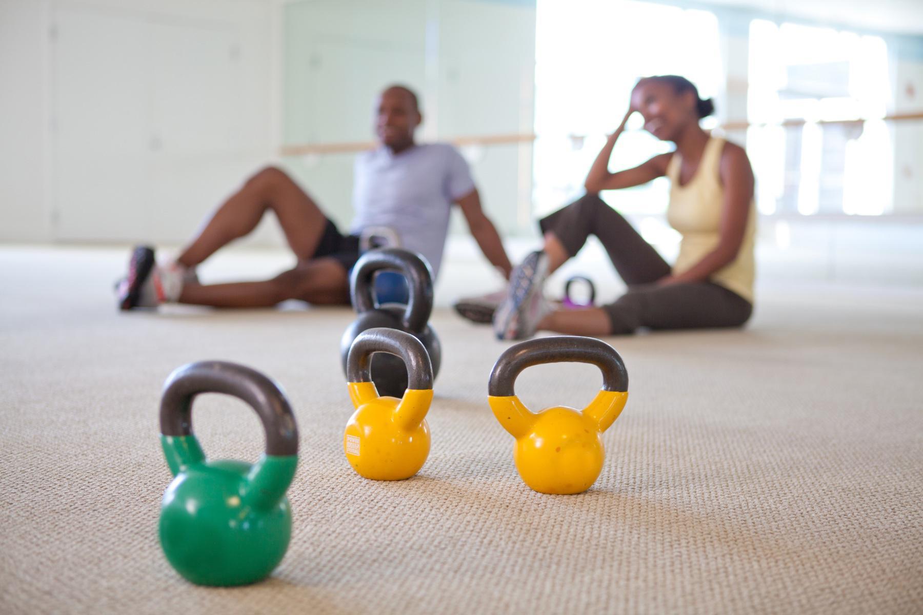 focus on bright yellow and green kettlebells sitting on workout studio floor with a blurry man and woman sitting on the ground in the background smiling