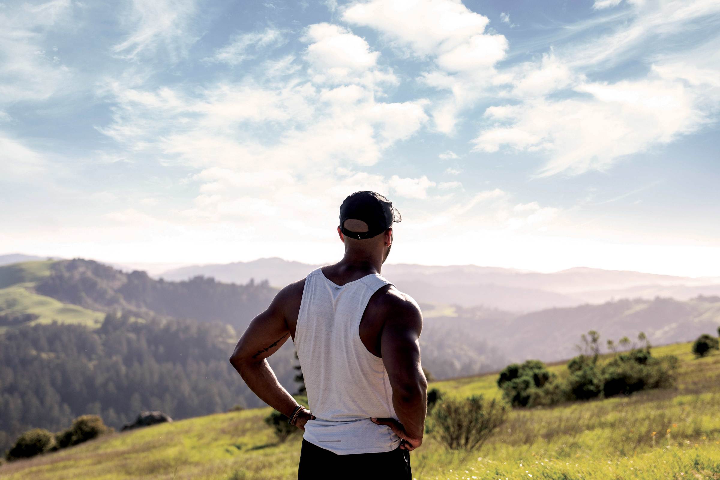 man's back as he stands with his hands on his hips overlooking a sunny valley