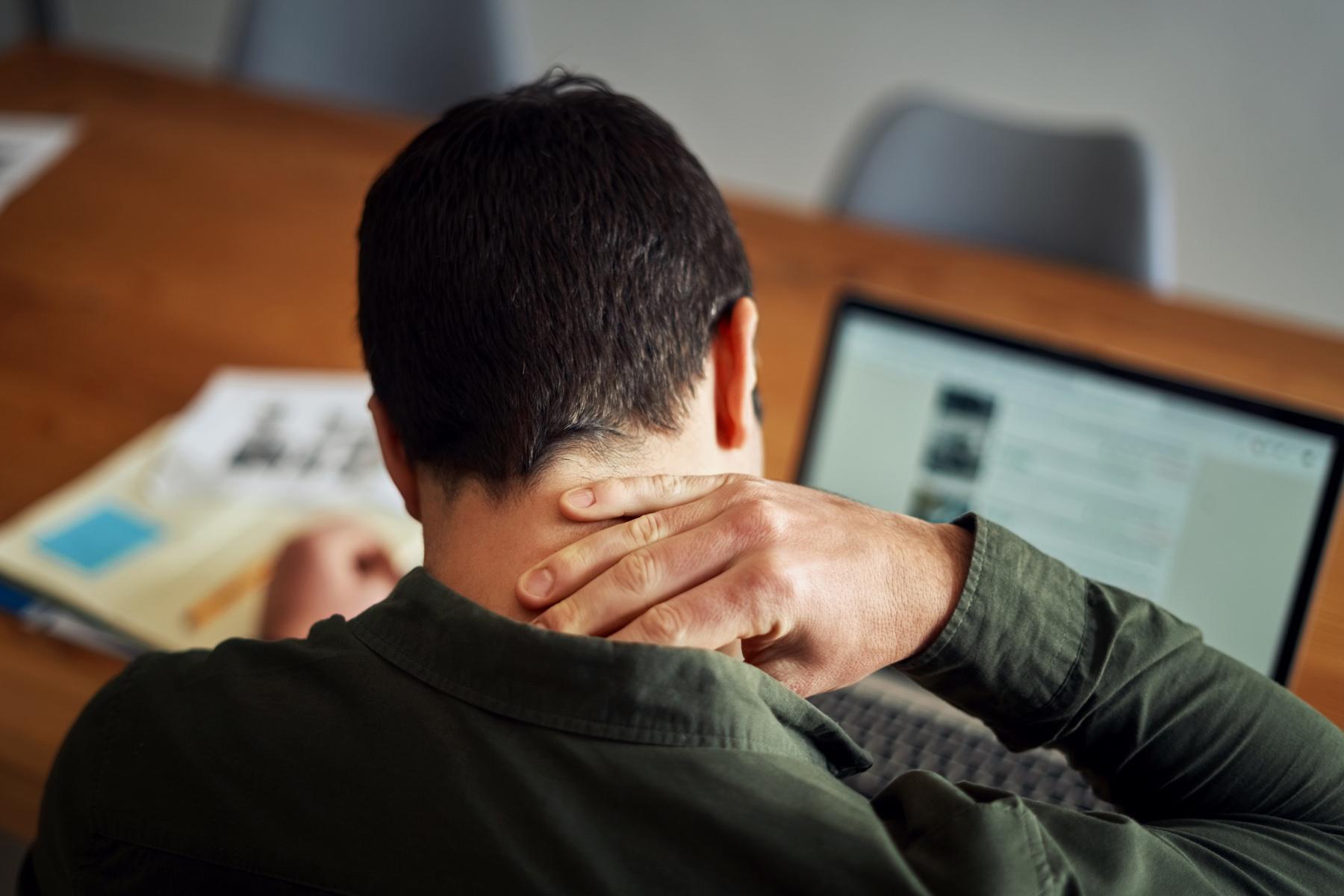man from behind rubbing his neck as he sits at laptop