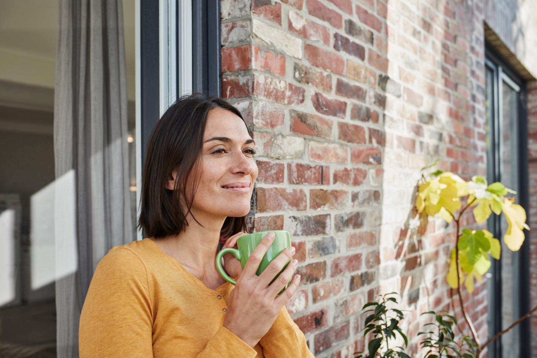 Dark-haired young woman standing outside with back against brick building smiling and holding a cup of tea.