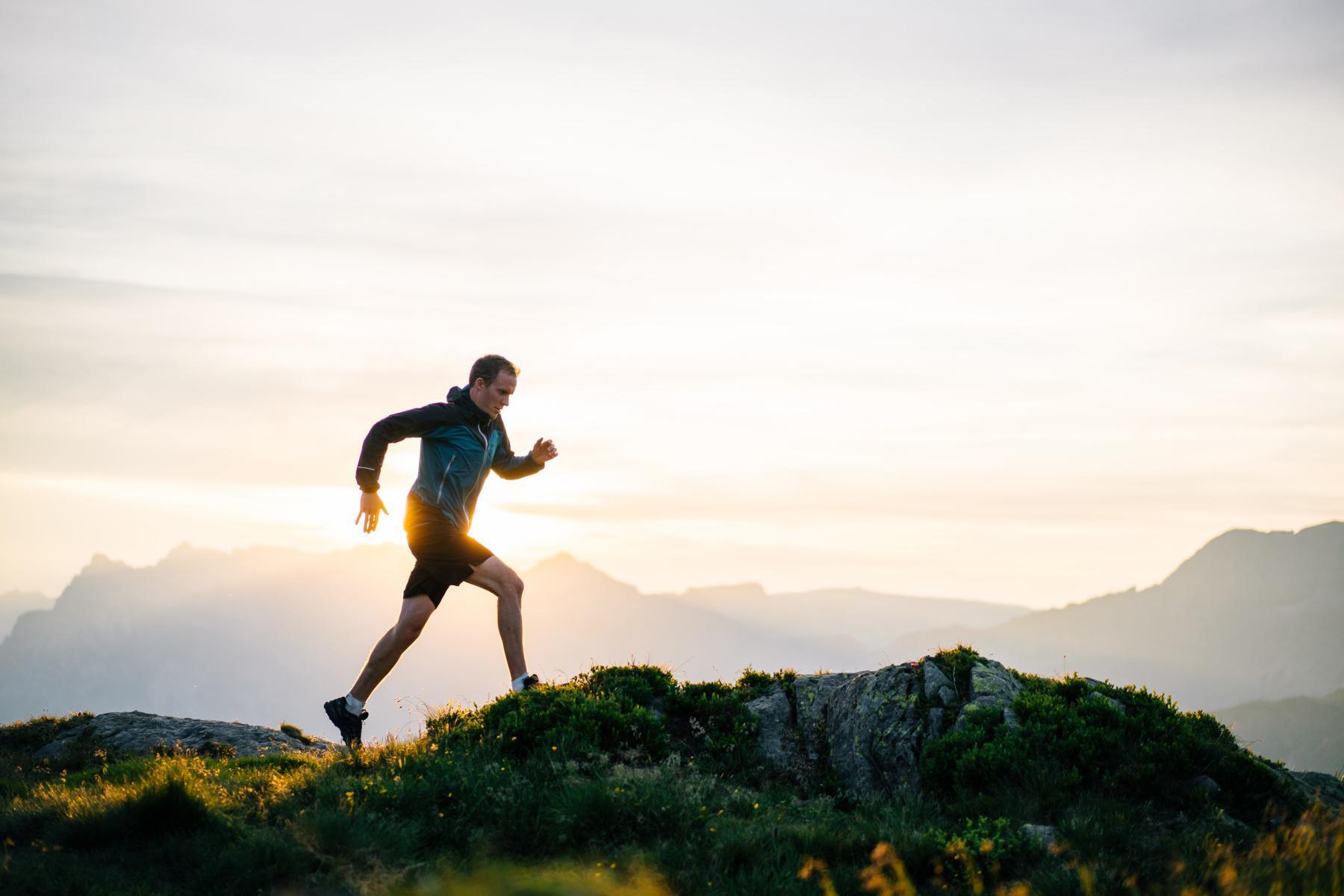 A male runner, silhouetted against the sky at sunset while running in the mountains on a steep trail.