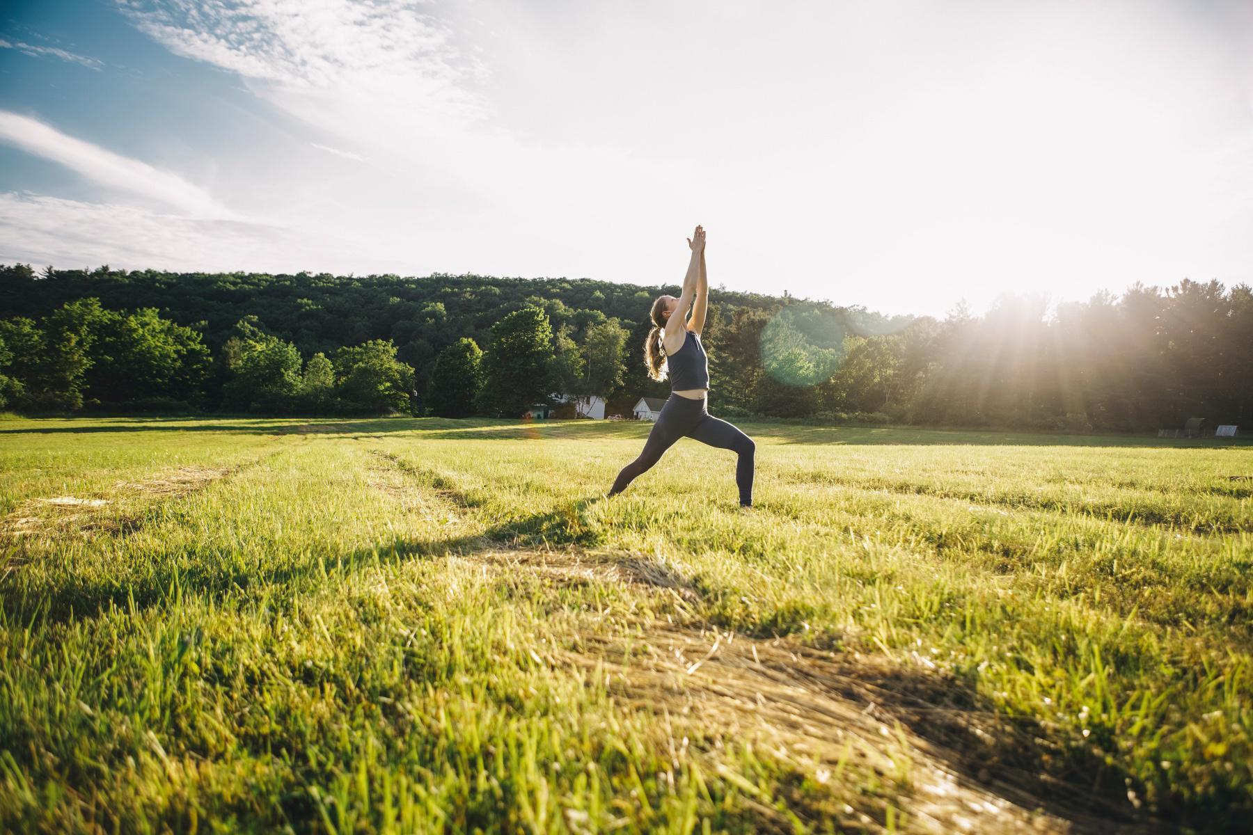 woman standing in a grassy field with the sun shining behind her as she does a warrior yoga pose
