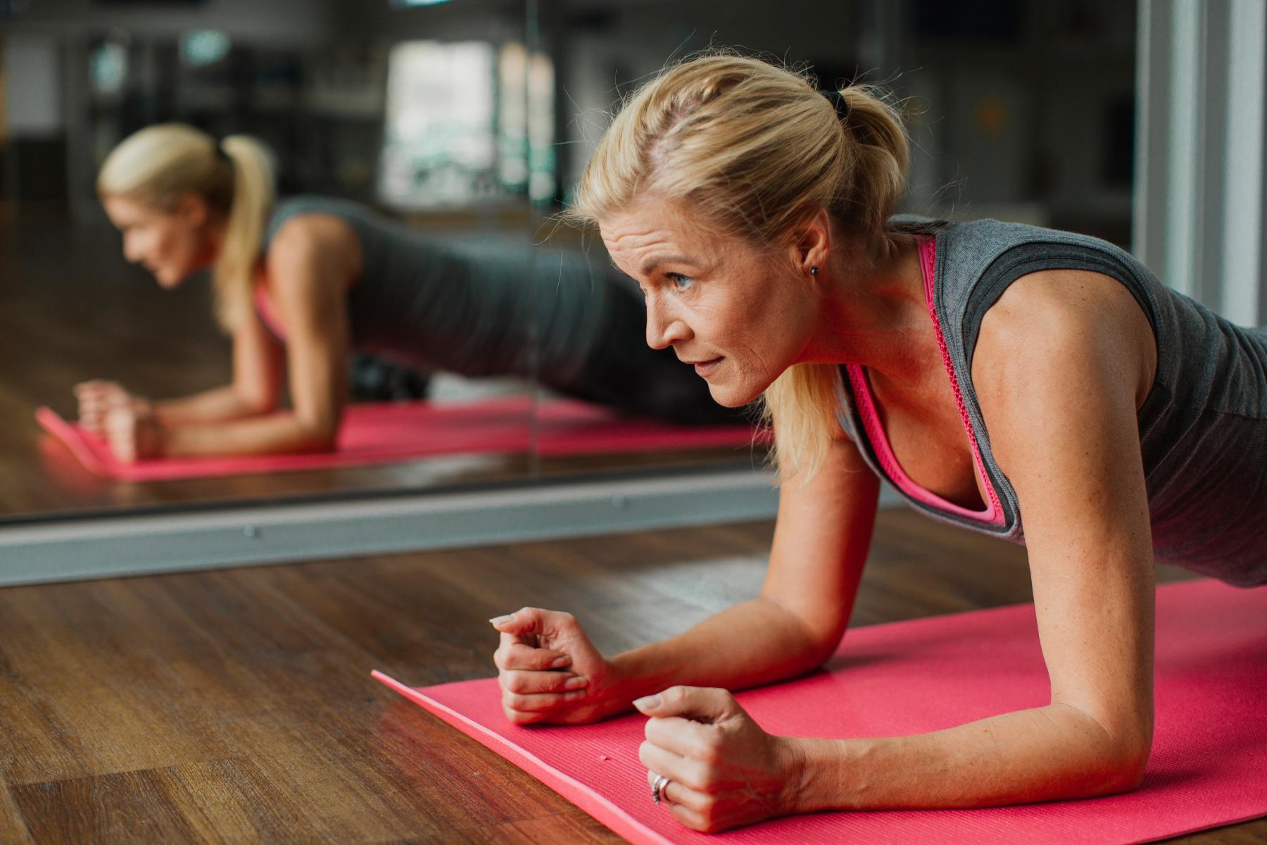 older woman in a plank position on a pink yoga mat in front of mirror