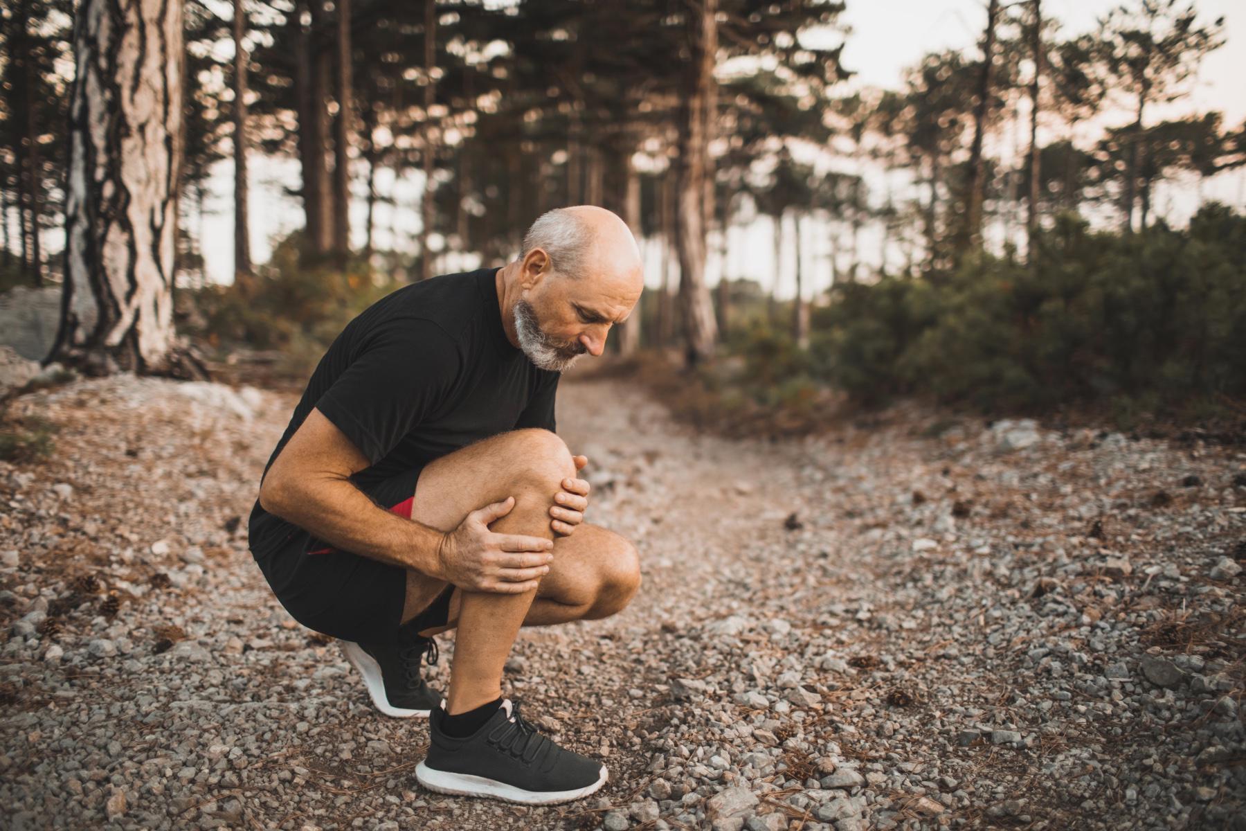 Older man bending down to hold his shin in pain as he is working out outside.