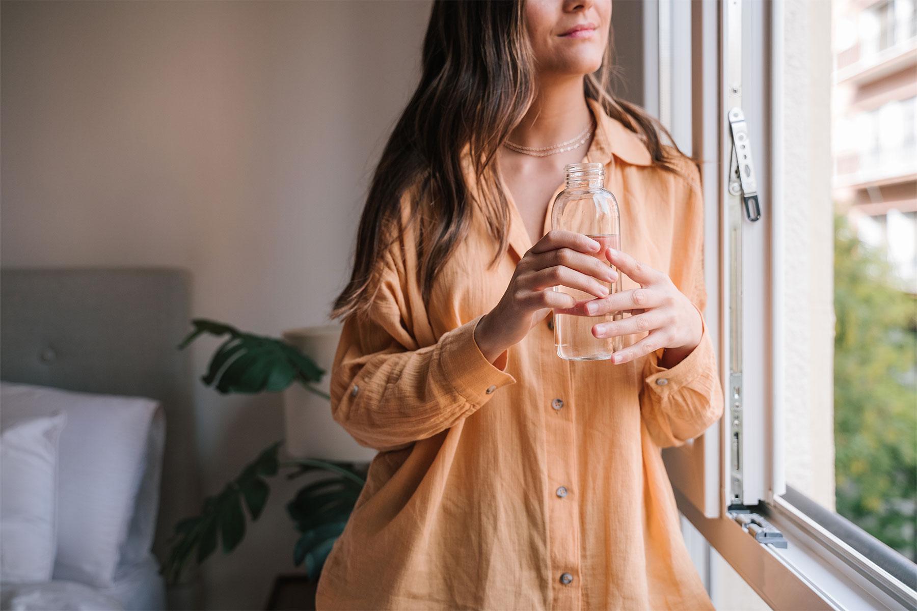 Woman leaning against window looking outside while holding a glass of water.