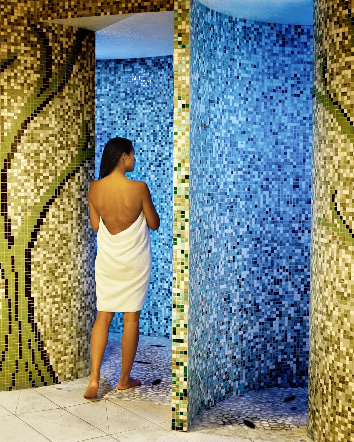 A young woman in a towel walking into the experiential rain entrance, which is an array of mosaic green tiles.