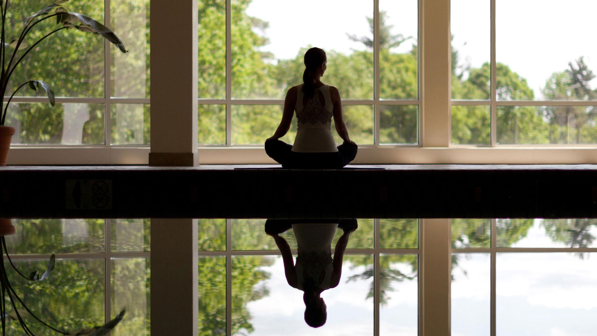 Woman practicing yoga poolside as she looks out the windows.