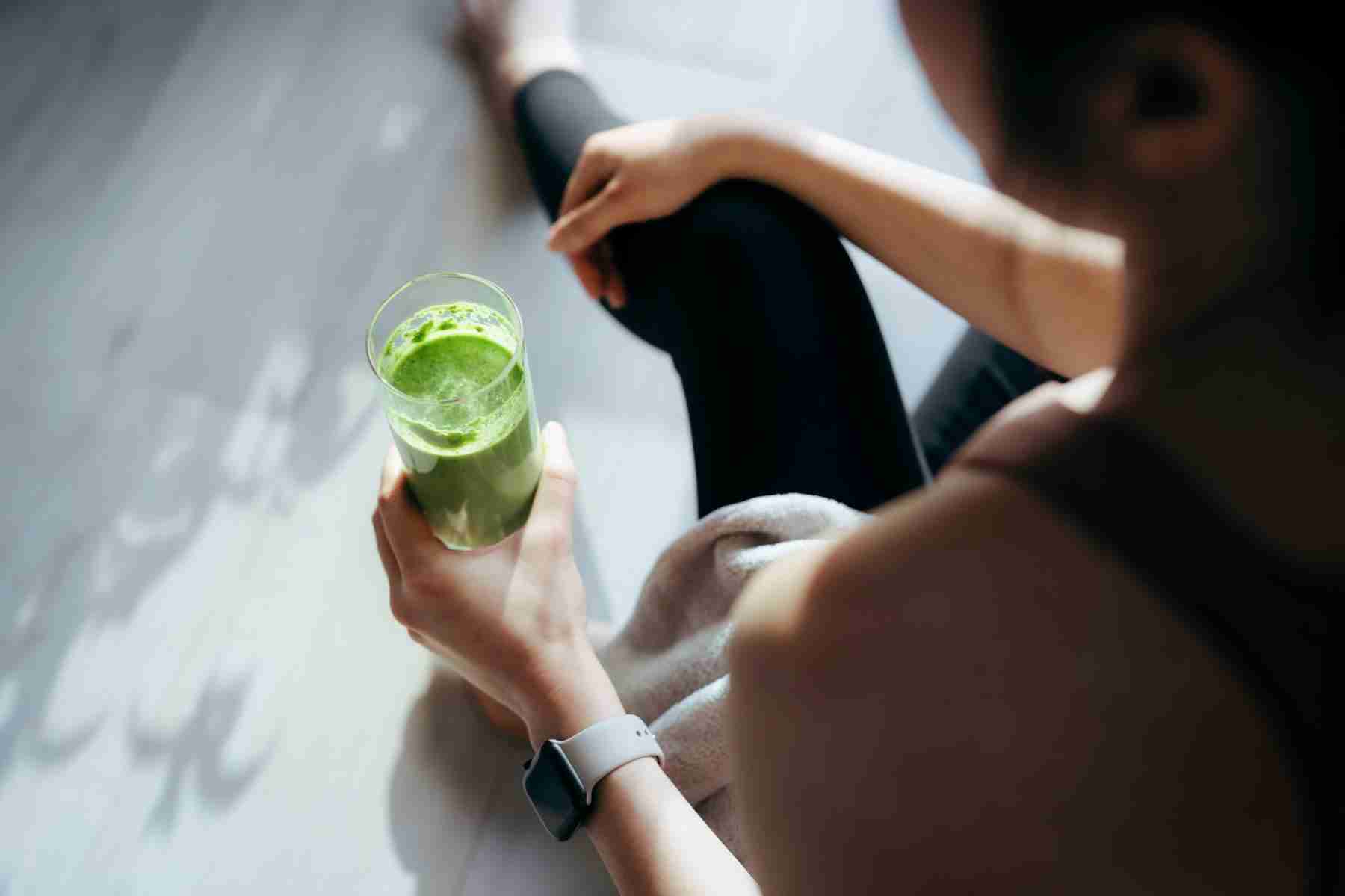 Woman sitting on the ground after working out drinking a green smoothie.