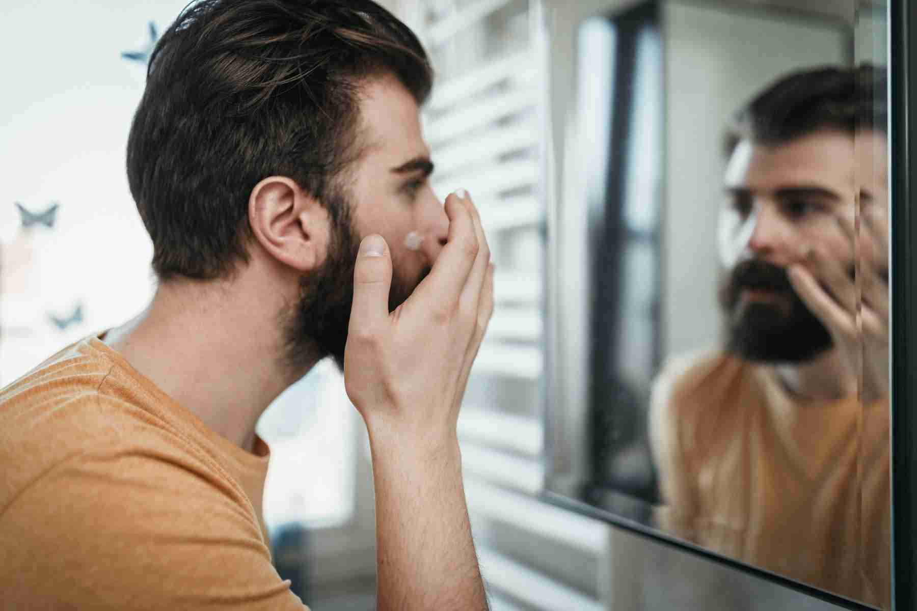 A man putting lotion on his face while looking into his bathroom mirror