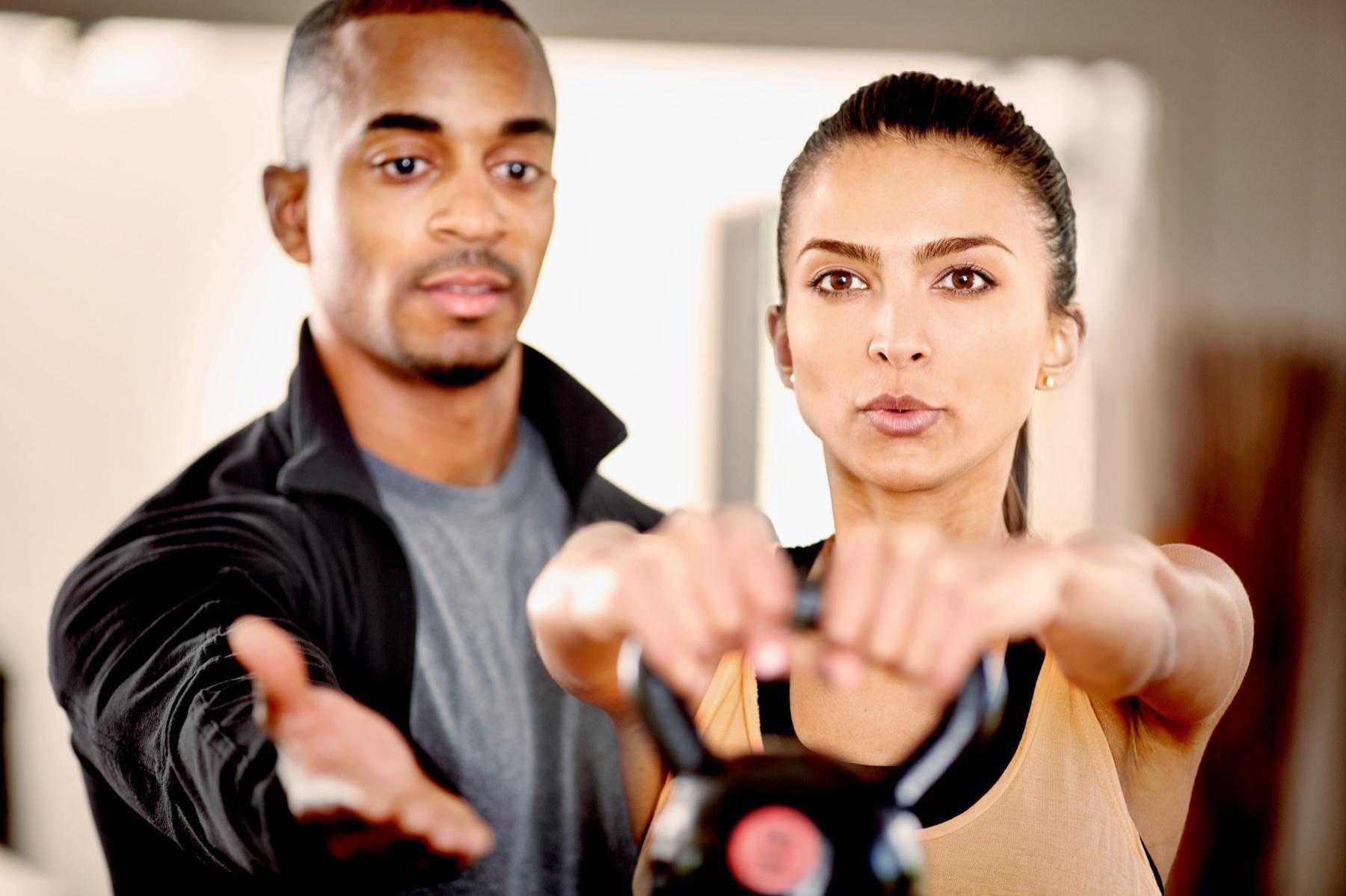 male personal trainer spotting woman as she raises a kettlebell in front of her
