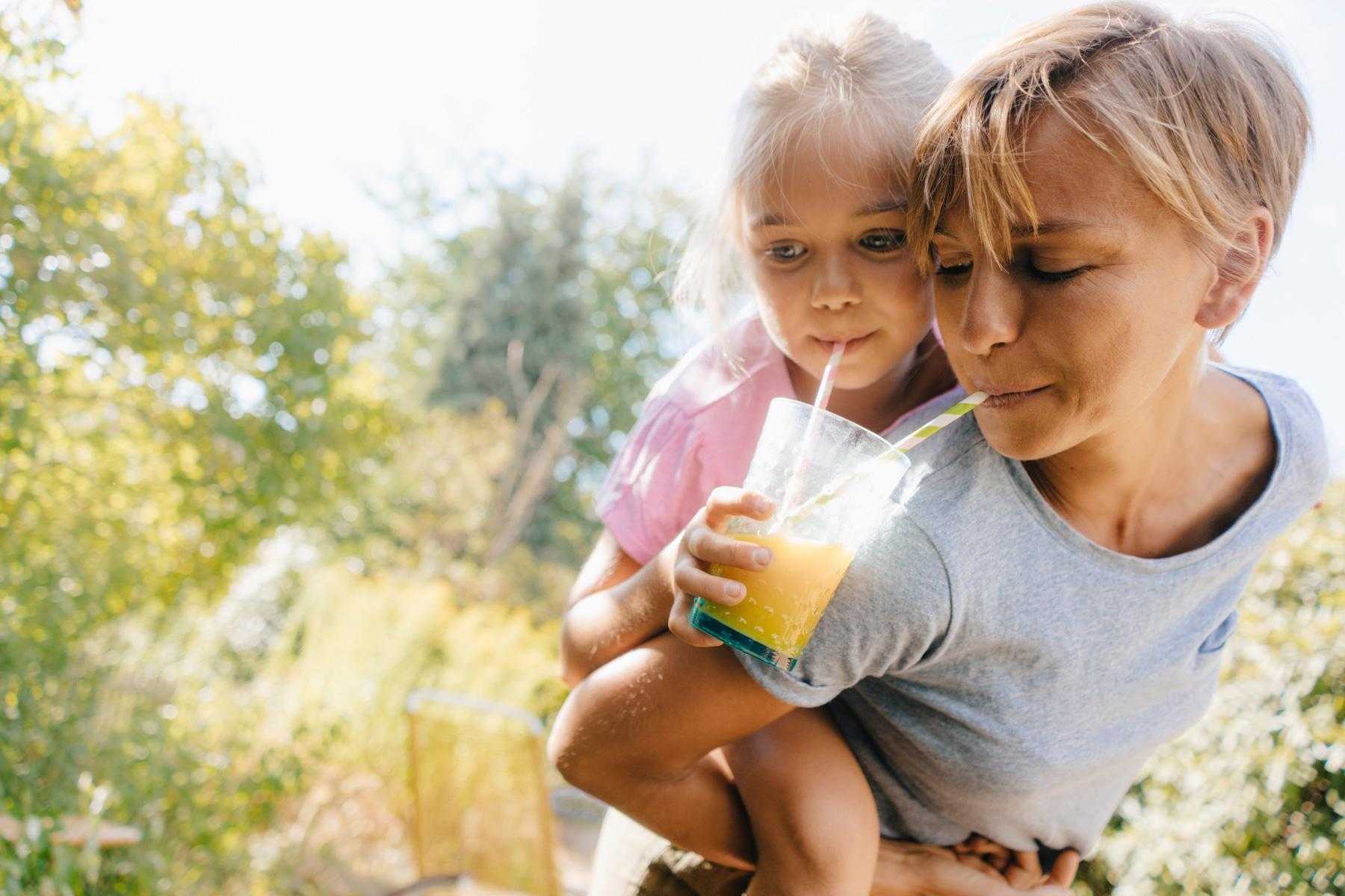 Mother with daughter on her back both drinking from same yellow smoothie with two straws.