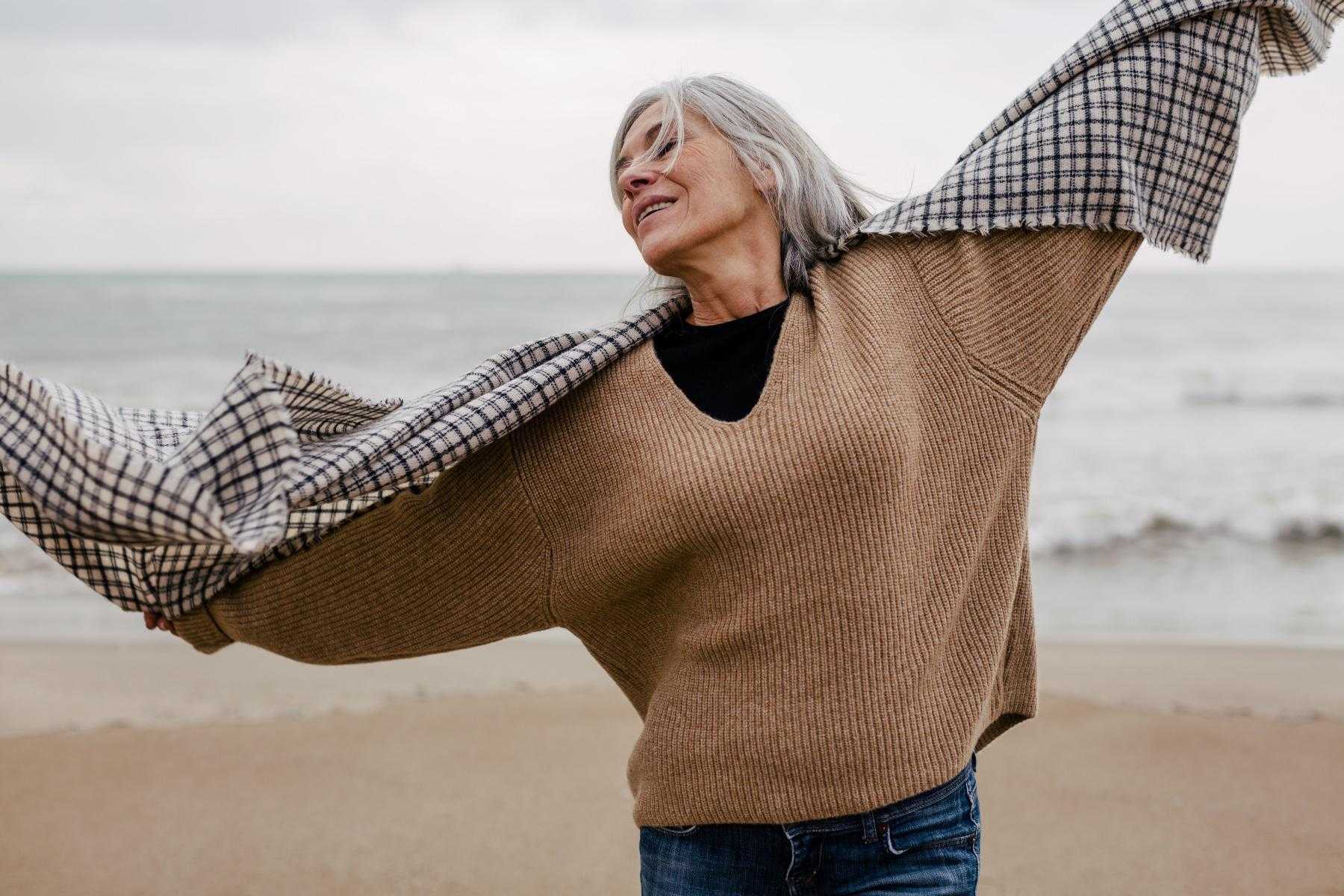 Older woman by ocean with scarf wrapped around her arms as she raises them up with her head back and eyes closed.