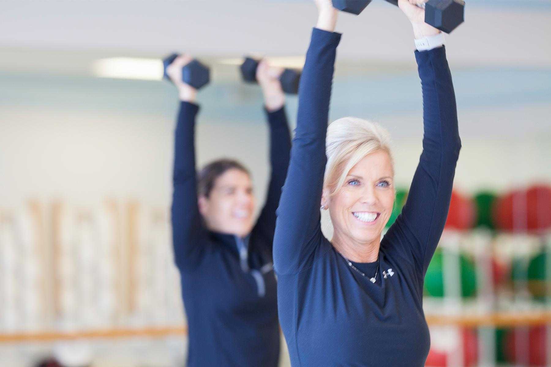 two women wearing all black in fitness room holding dumbbells above their head during class