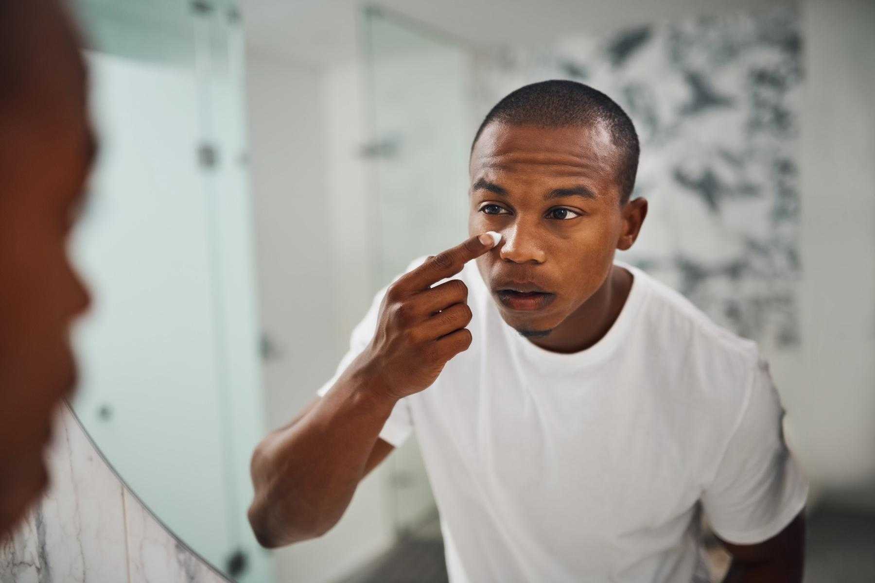 Young man standing in front of his bathroom mirror at home applying lotion to his face.