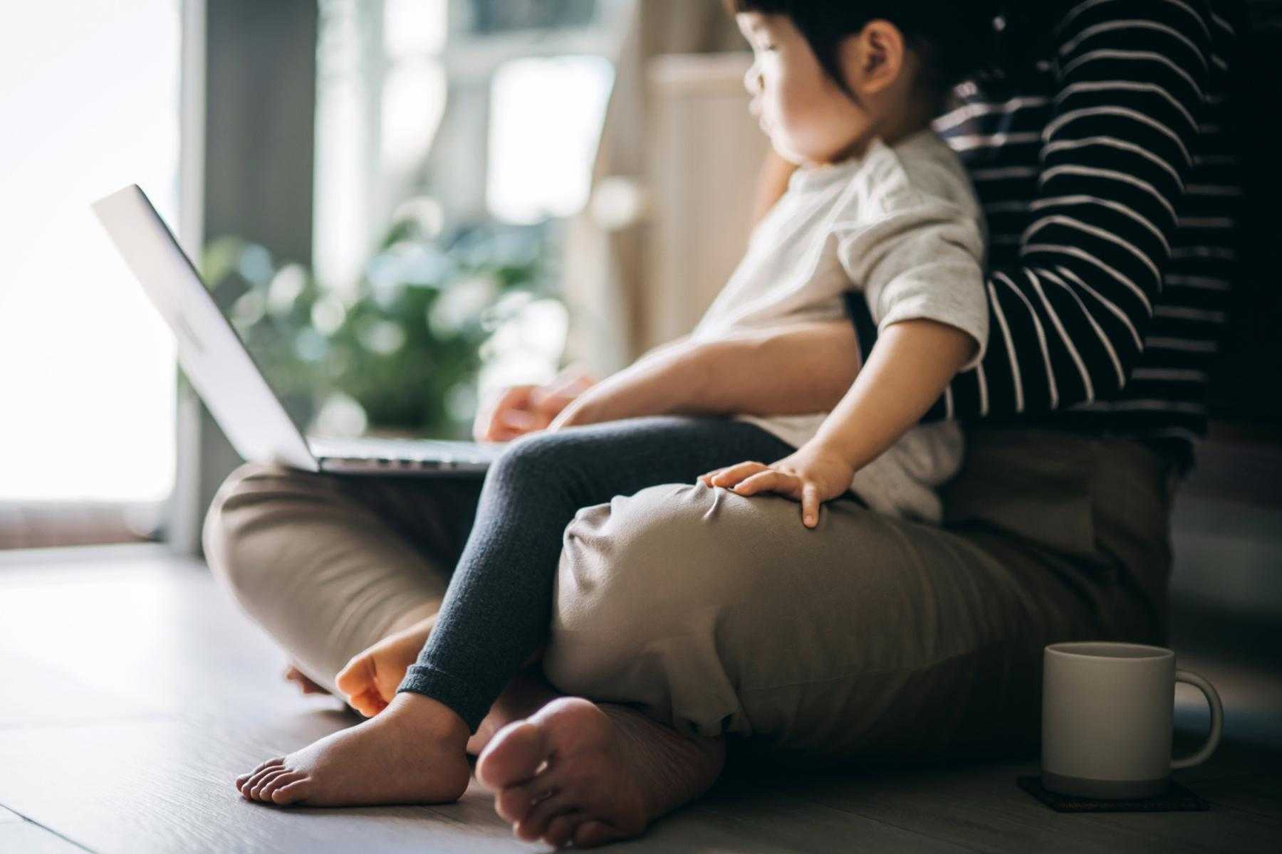 Woman sitting on ground working on laptop with toddler sitting in lap.