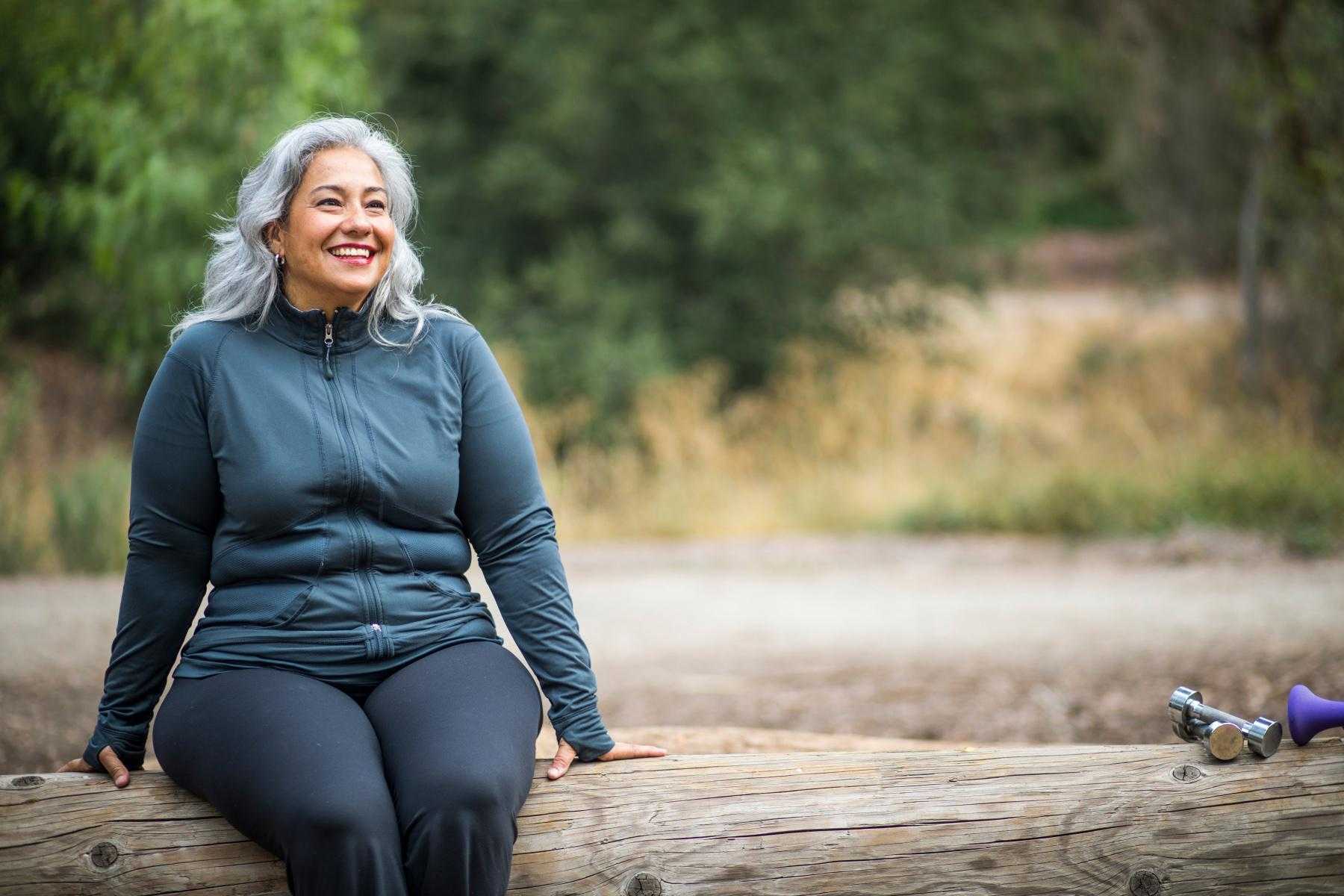 Woman smiling and looking forward while sitting on a log outside.