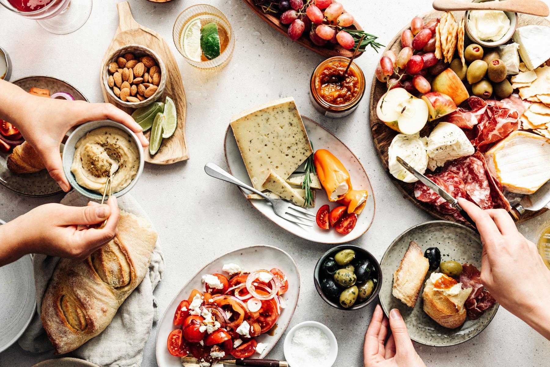 overhead table view of hands serving from plates of mediterranean food including cheese and olives
