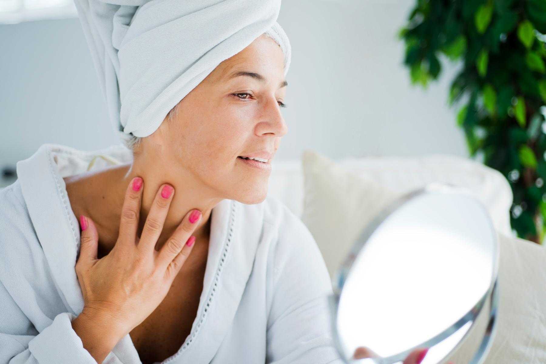 A woman in a robe and her hair in a towel looking at her neck in a handheld mirror.