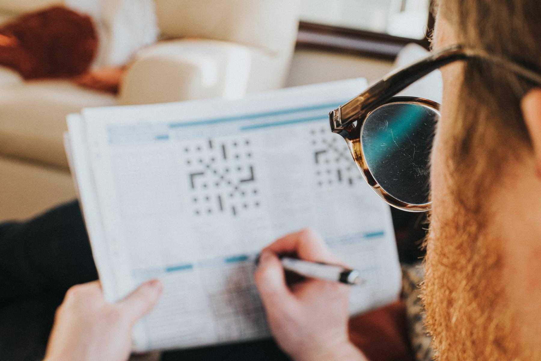 Looking over the shoulder of a man with glasses on doing a crossword puzzle in the newspaper while sitting on a couch.