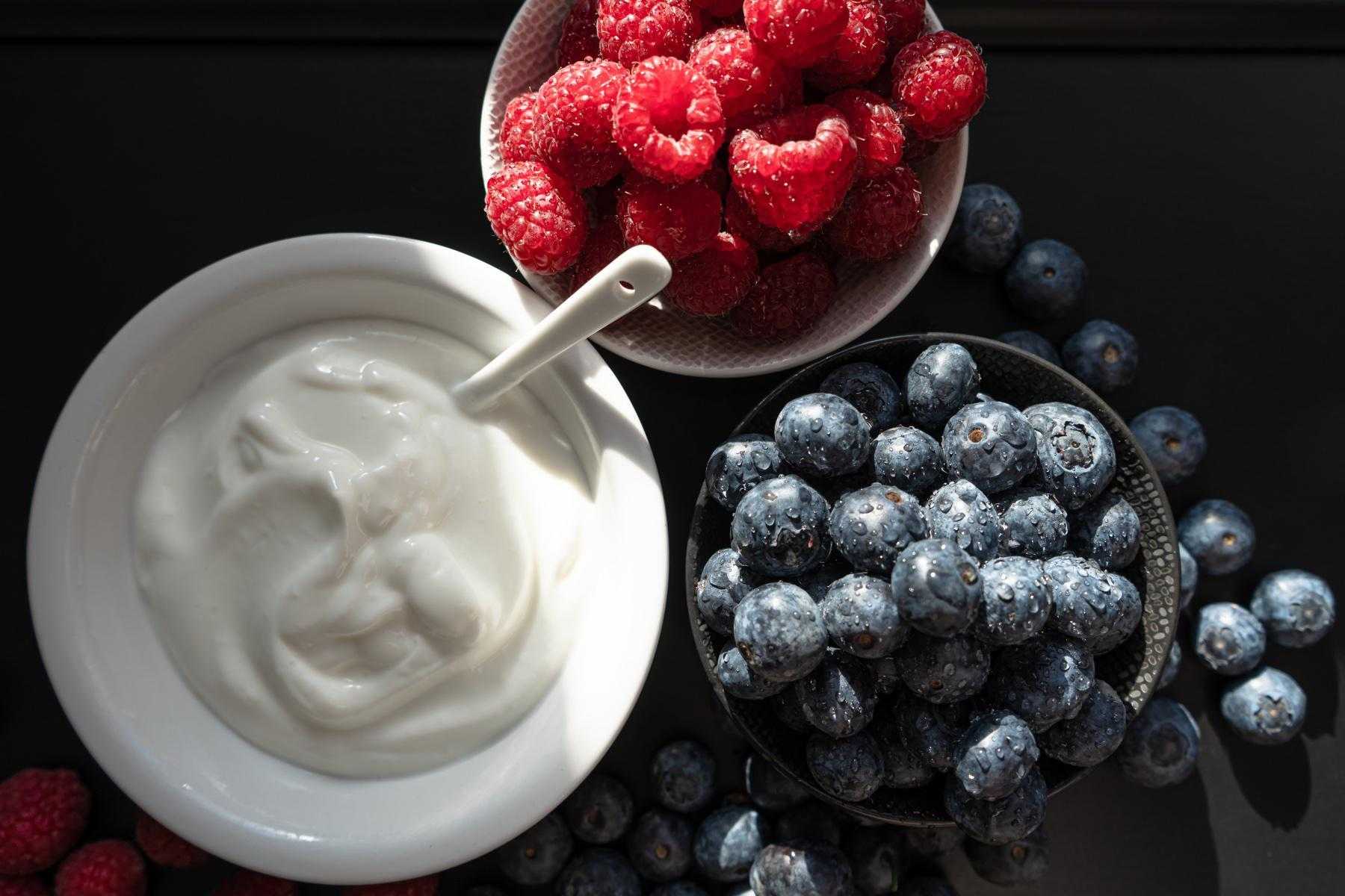 Close-up overhead shot of a bowl of yogurt next to a bowl of raspberries and a bowl of blueberries.