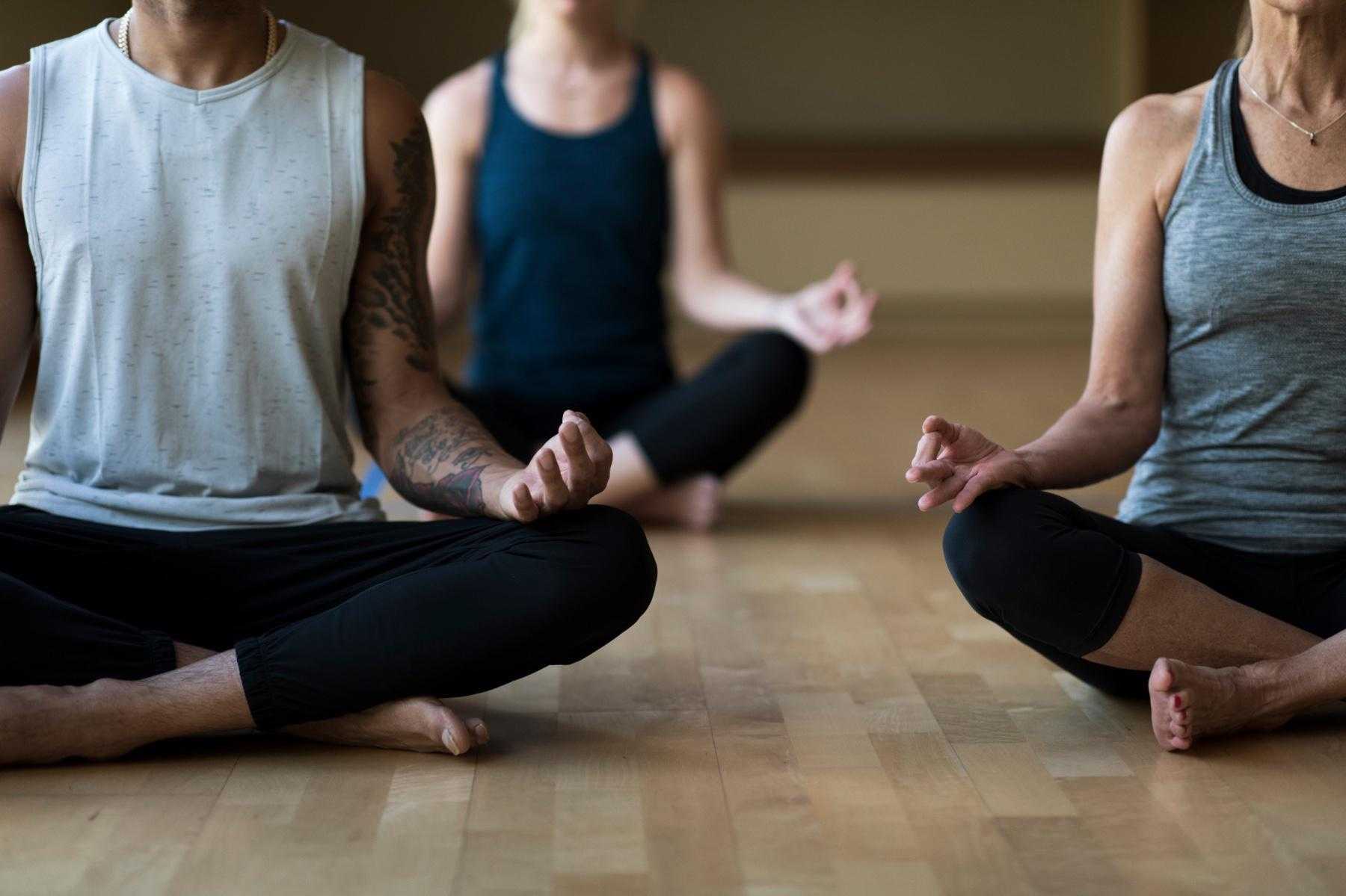 Three people shown from the neck down in a yoga studio sitting in a cross-legged meditation pose.