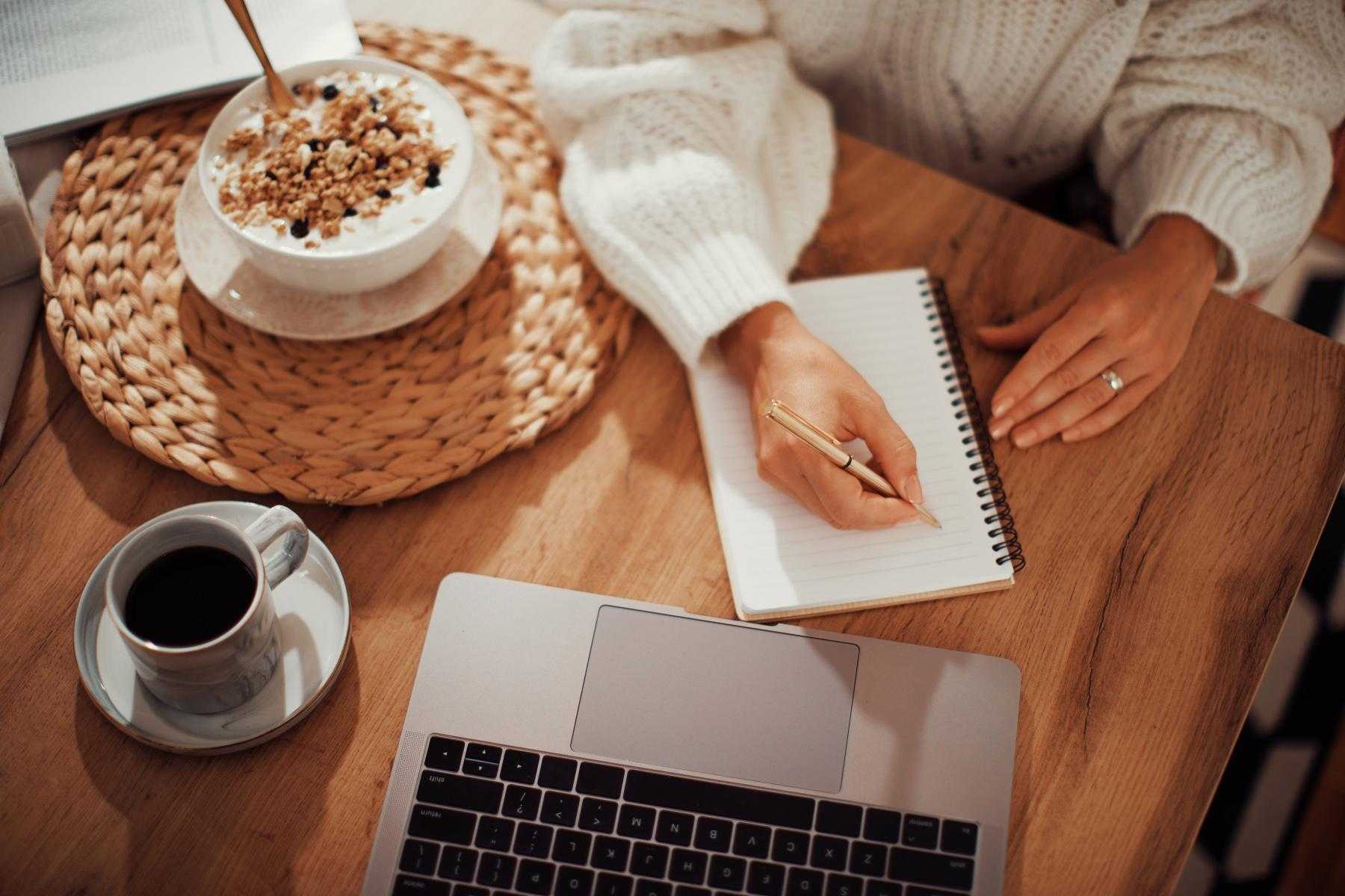 overhead view of woman writing in notebook next to laptop with bowl of yogurt next to her