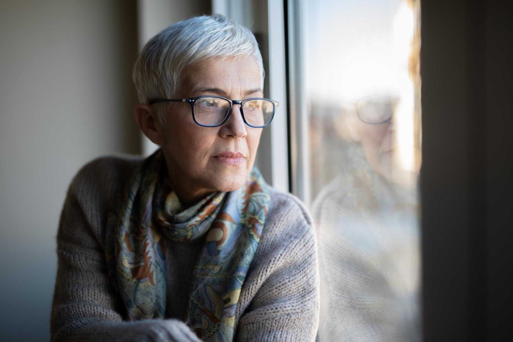 Older woman with glasses sitting up against window looking outside.