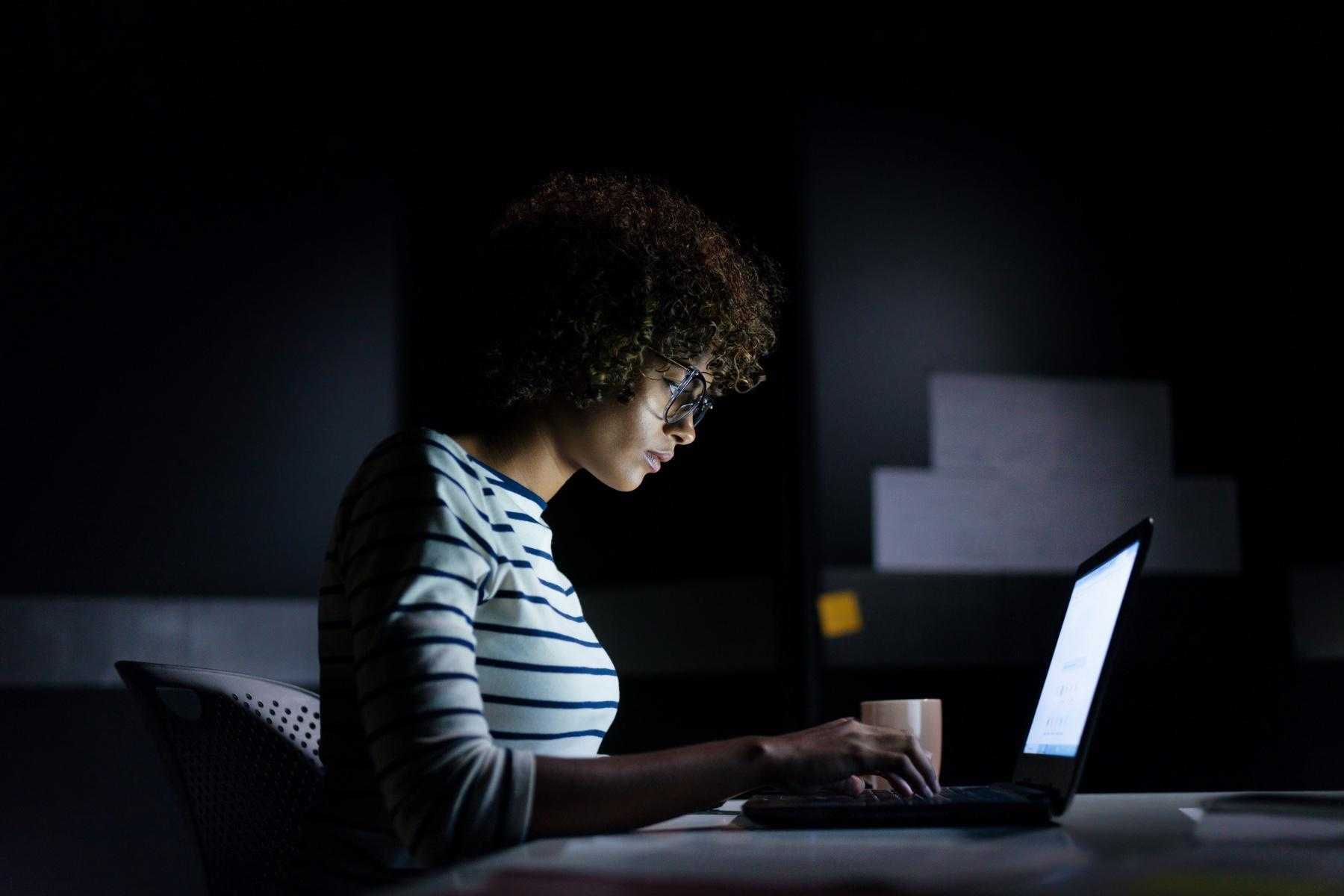 Side view of woman sitting at a table in a dark room looking at her laptop screen.
