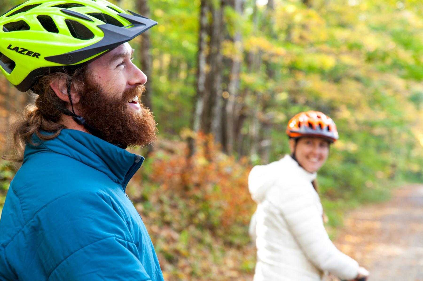 close-up of man with beard smiling with bike helmet on, while woman in back looks back and smiles on her bike