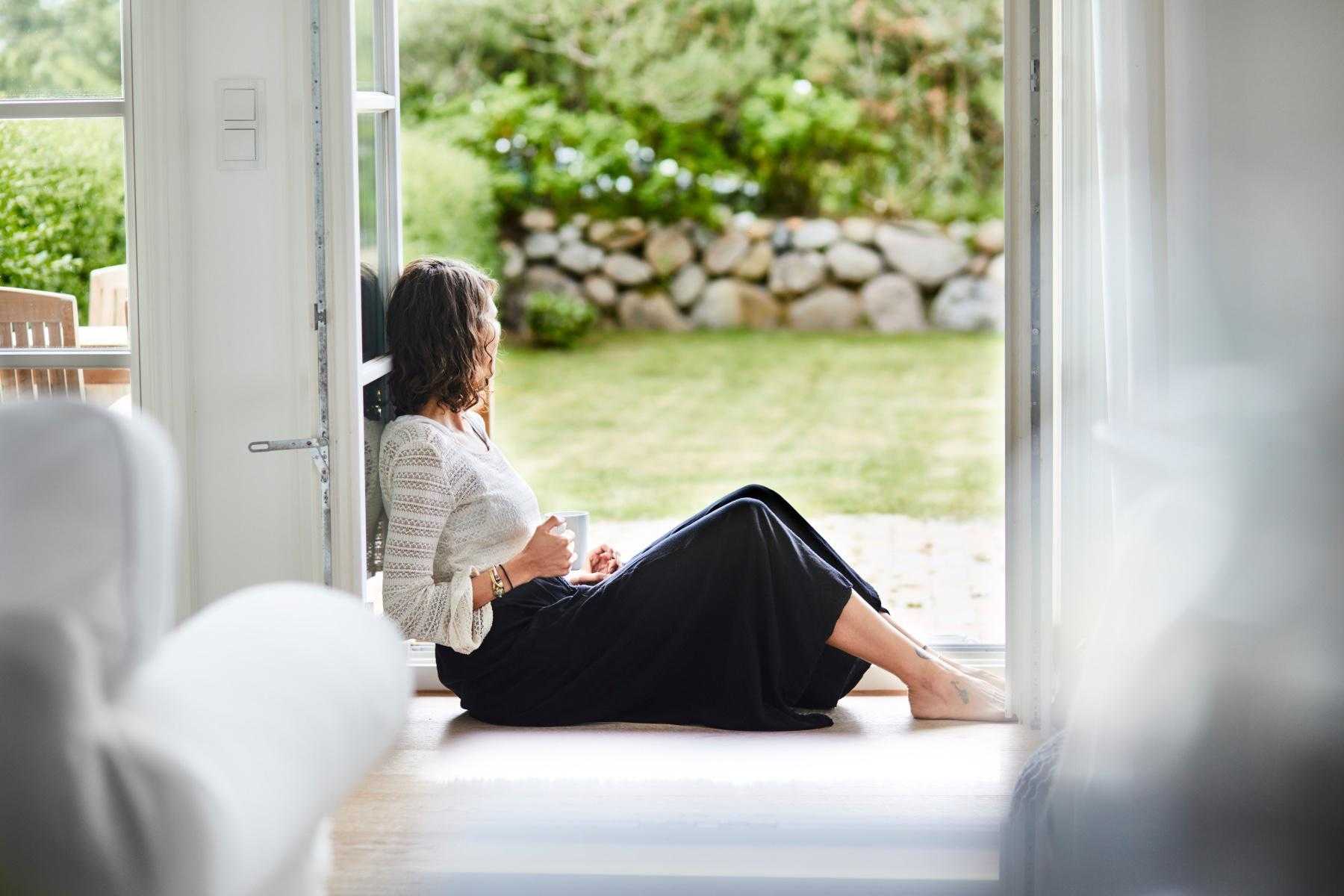 Woman holding a coffee cup while sitting on the floor in a doorway looking outside at nature.