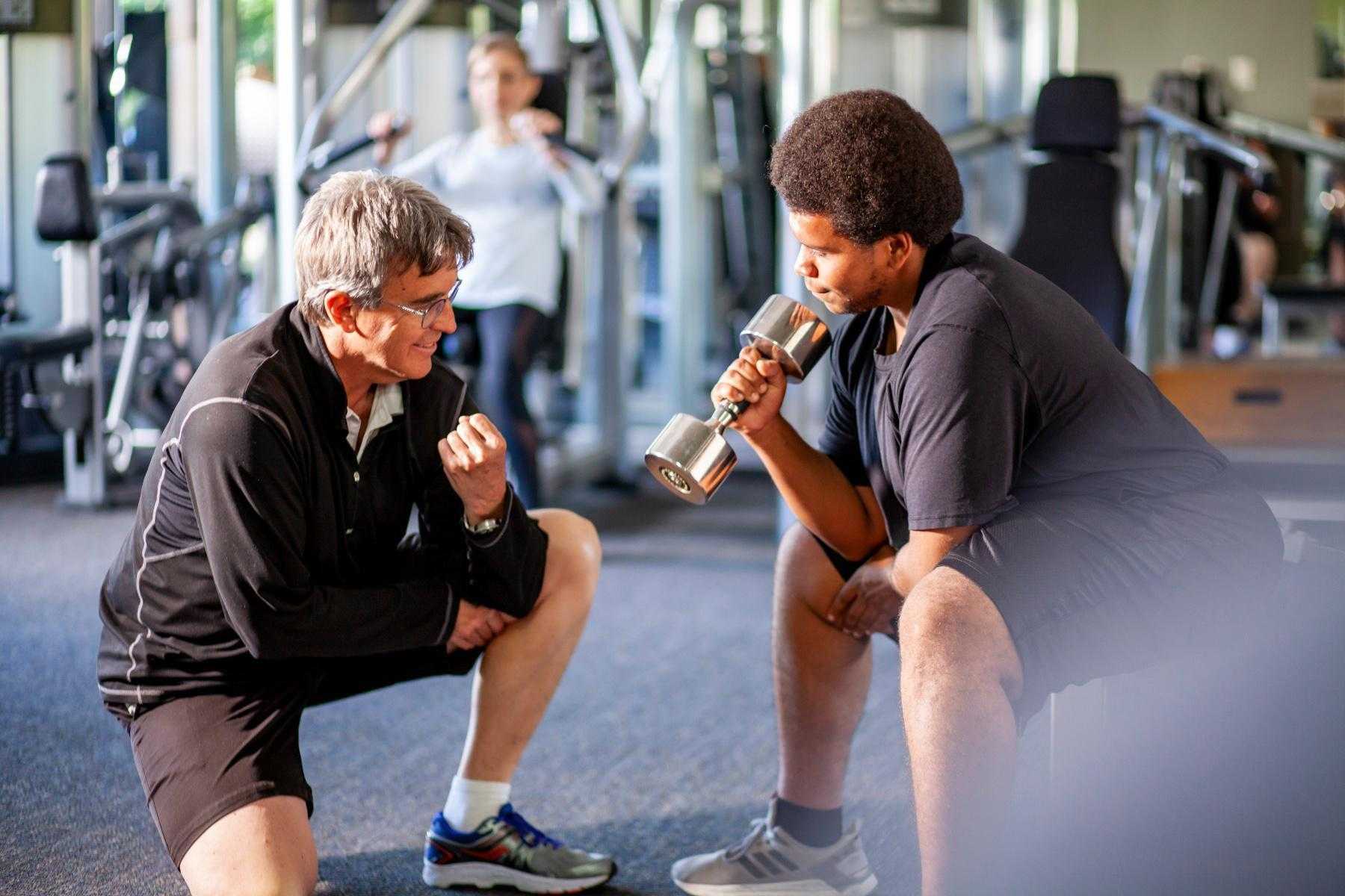 man kneeling on the ground showing another man how to perform a bicep curl